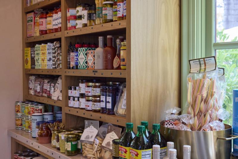 A well-organized oak built-in shelving unit in a specialty grocery or gourmet food shop, filled with jars, bottles, broths, and packaged goods, with natural light coming through a green-trimmed window.