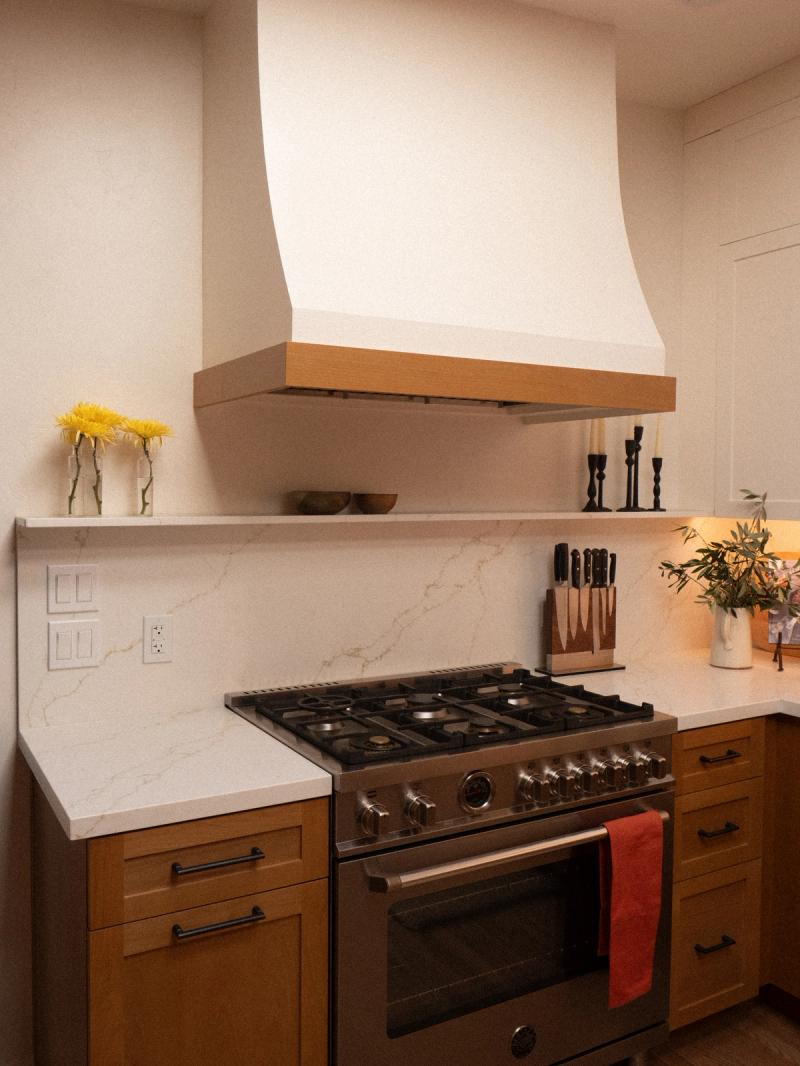 A custom kitchen featuring warm oak cabinetry with black hardware, white quartz countertops and backsplash, a stainless steel professional range, and a curved plaster range hood with a wood trim accent.