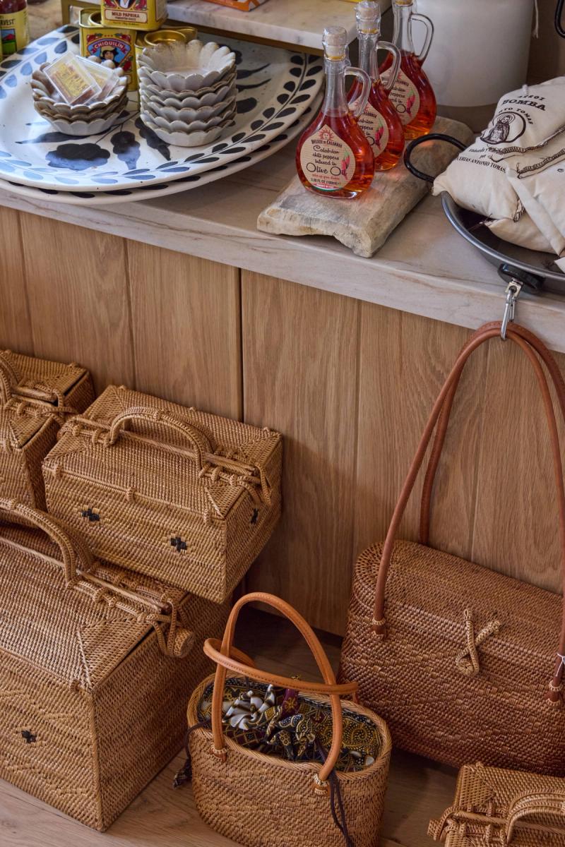A close-up of light oak wood cabinetry and countertop displaying decorative ceramic plates, olive oil bottles, and woven rattan baskets and bags arranged on the floor below.