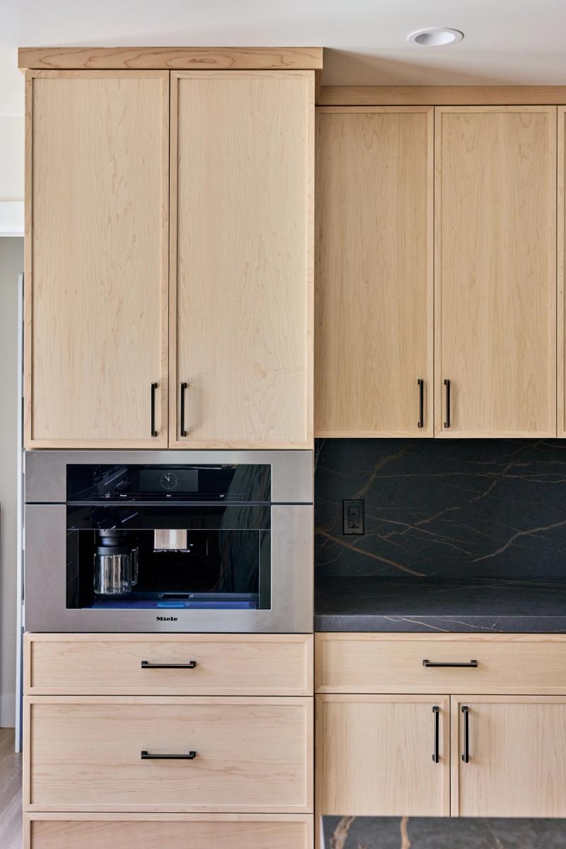 Custom light maple kitchen cabinetry with black matte hardware, featuring a built-in Miele coffee machine and dark stone backsplash and countertops.