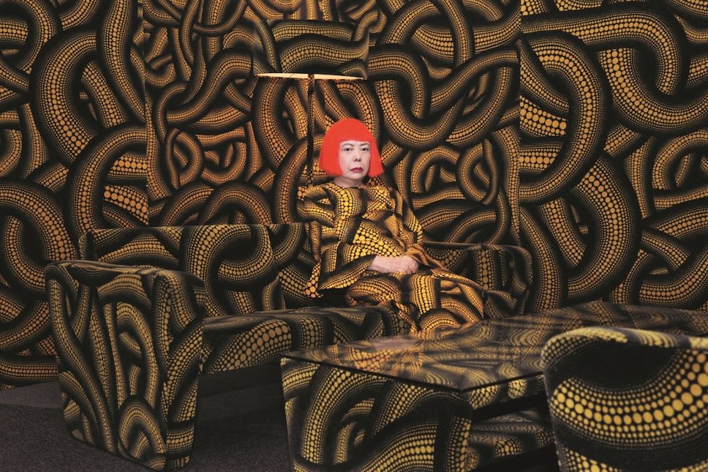 Yayoi Kusama in a red wig, seated in her installation Yellow Tree / Living Room, every surface covered in yellow and black tangled patterns.