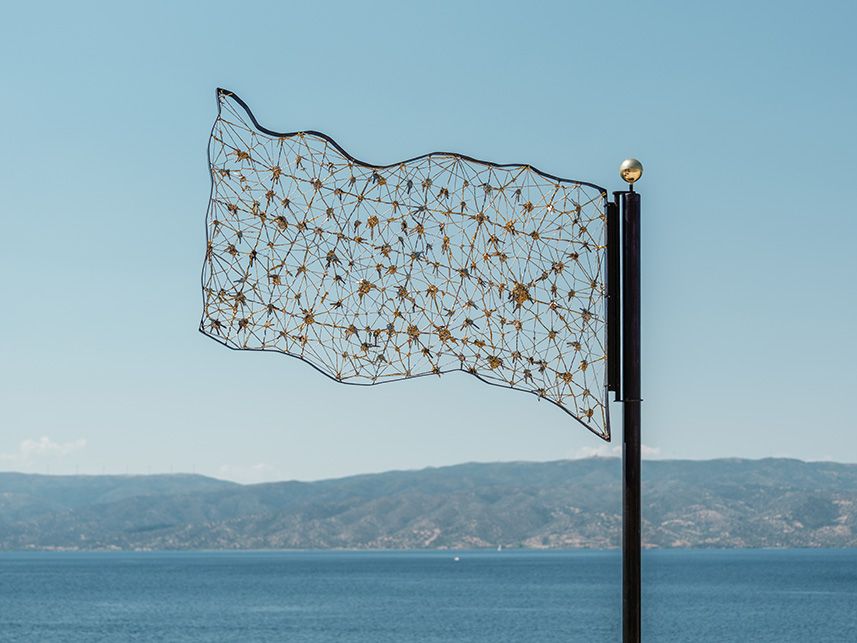 A flag woven from keys and metal mesh on a dark pole above the sea at Hydra.