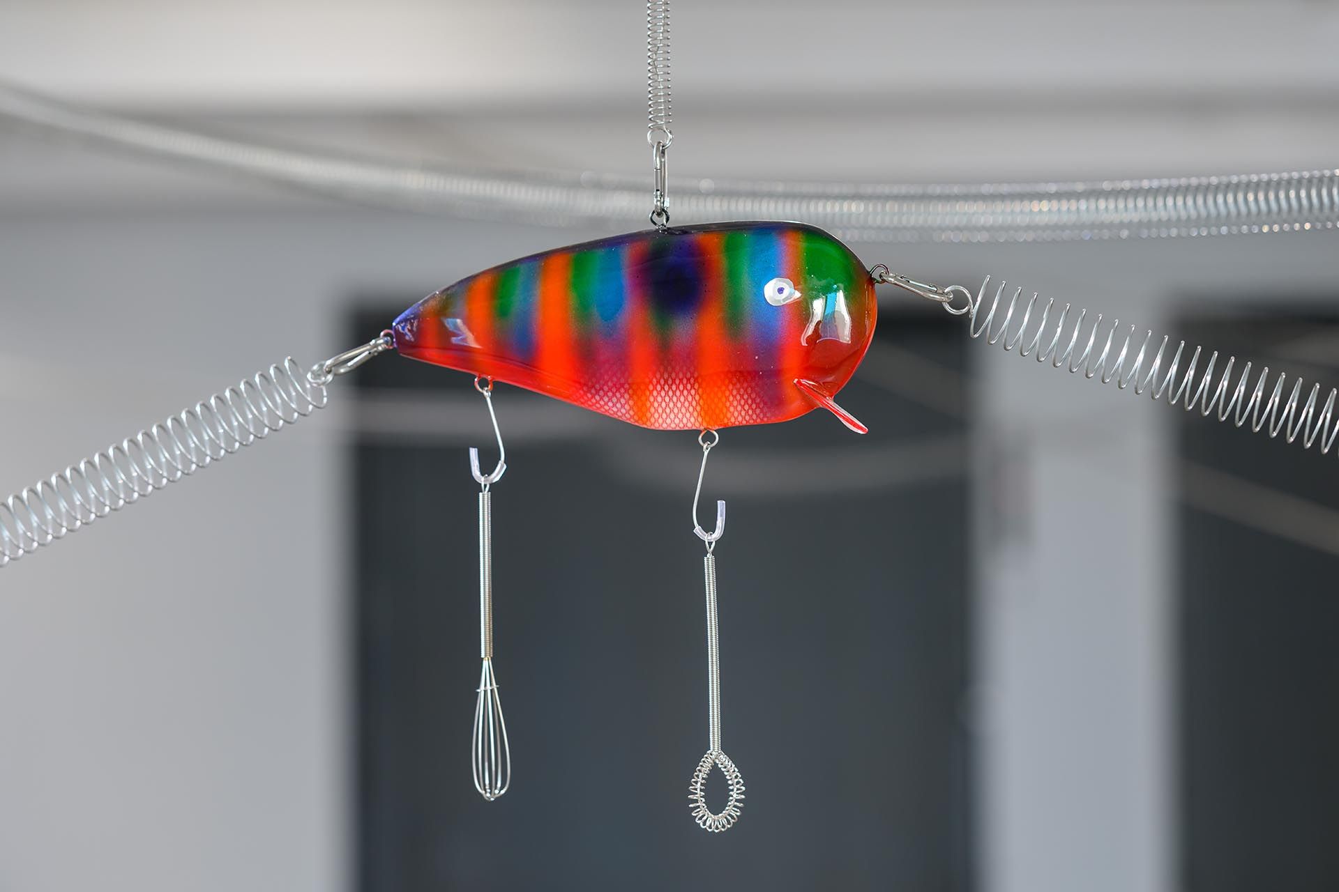 Detail of Aki Sasamoto's Sounding Lines: an oversized painted fishing lure suspended on metal springs, with two small whisks hanging beneath it.