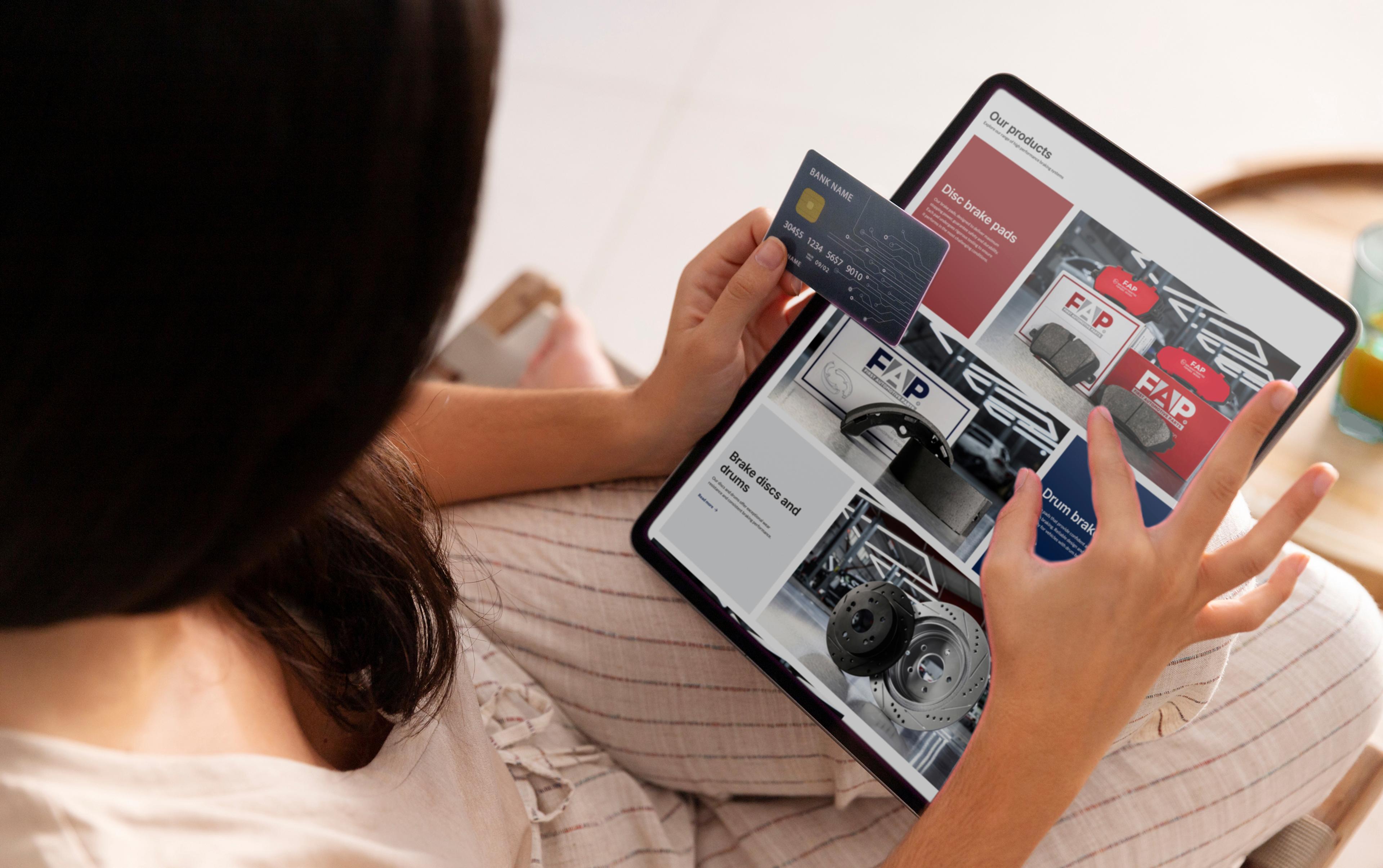 Человек держит в руках банковскую карту и использует планшет, на котором отображаются страницы товаров с тормозными колодками, тормозными дисками и упаковкой бренда FAP.