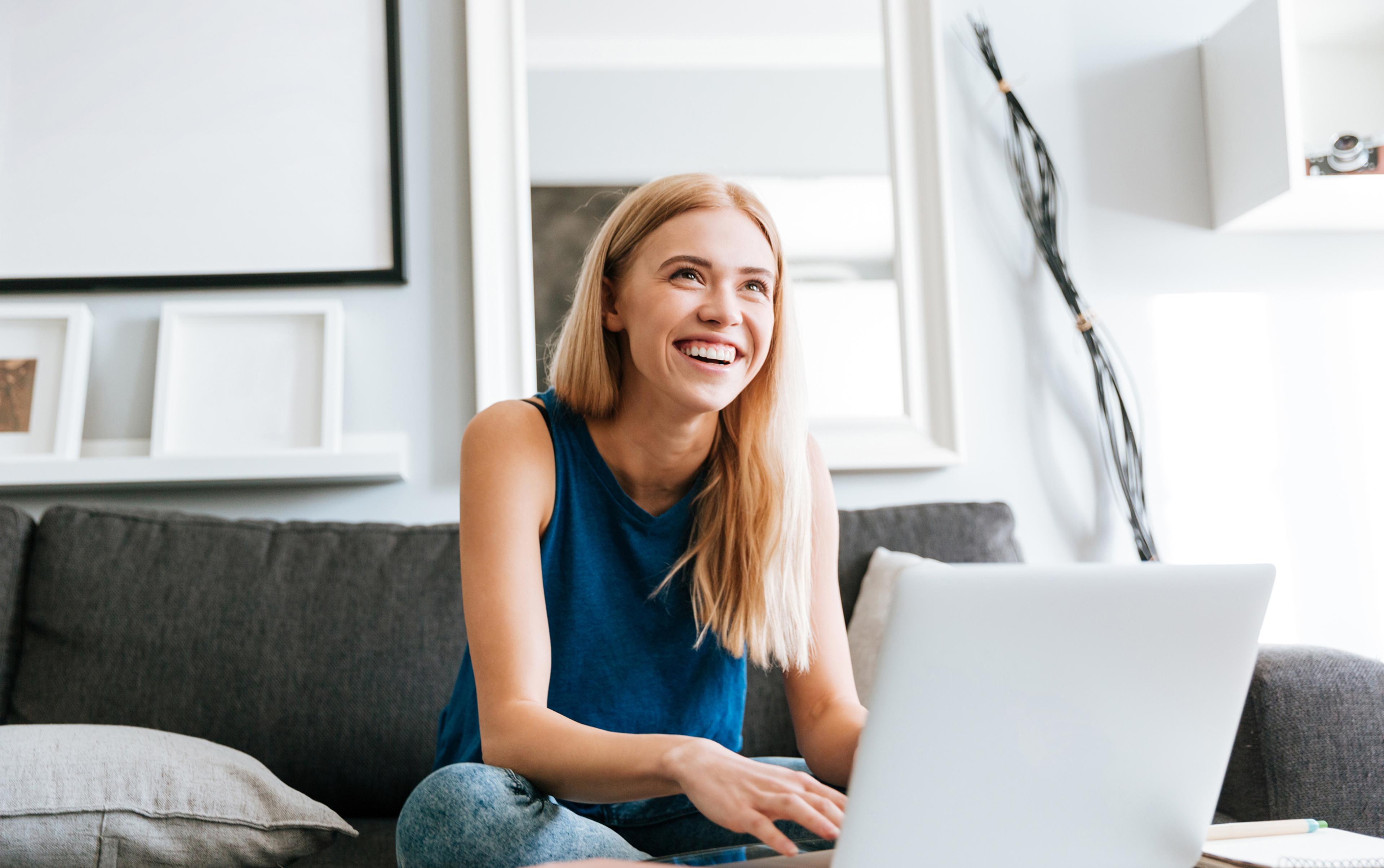 Junge Frau sitzt auf grauer Couch, lächelnd und Laptop nutzend, umgeben von heller Einrichtung.