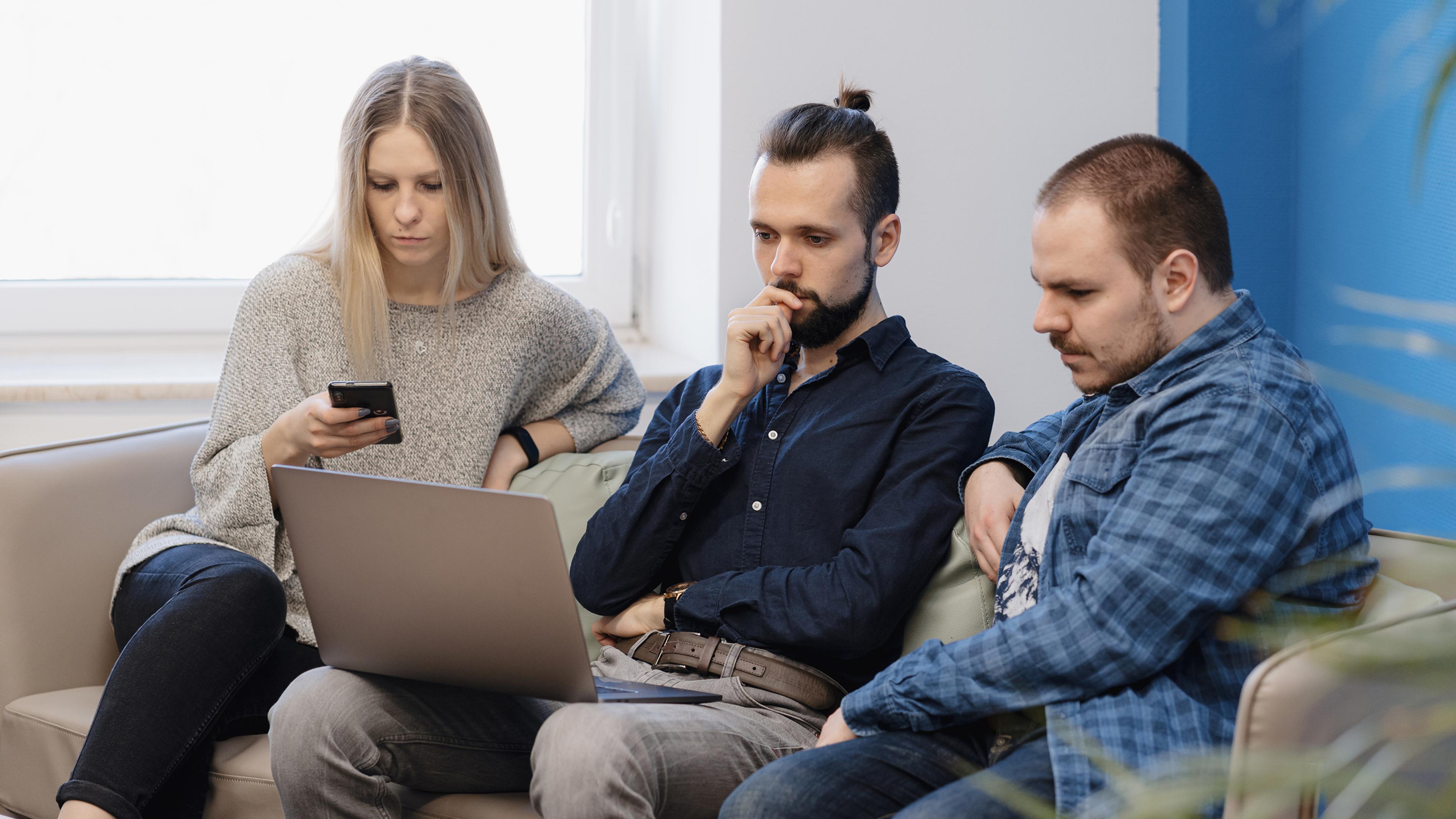 Drei Menschen sitzen auf einem Sofa und schauen auf einen Laptop, einer nutzt ein Handy.