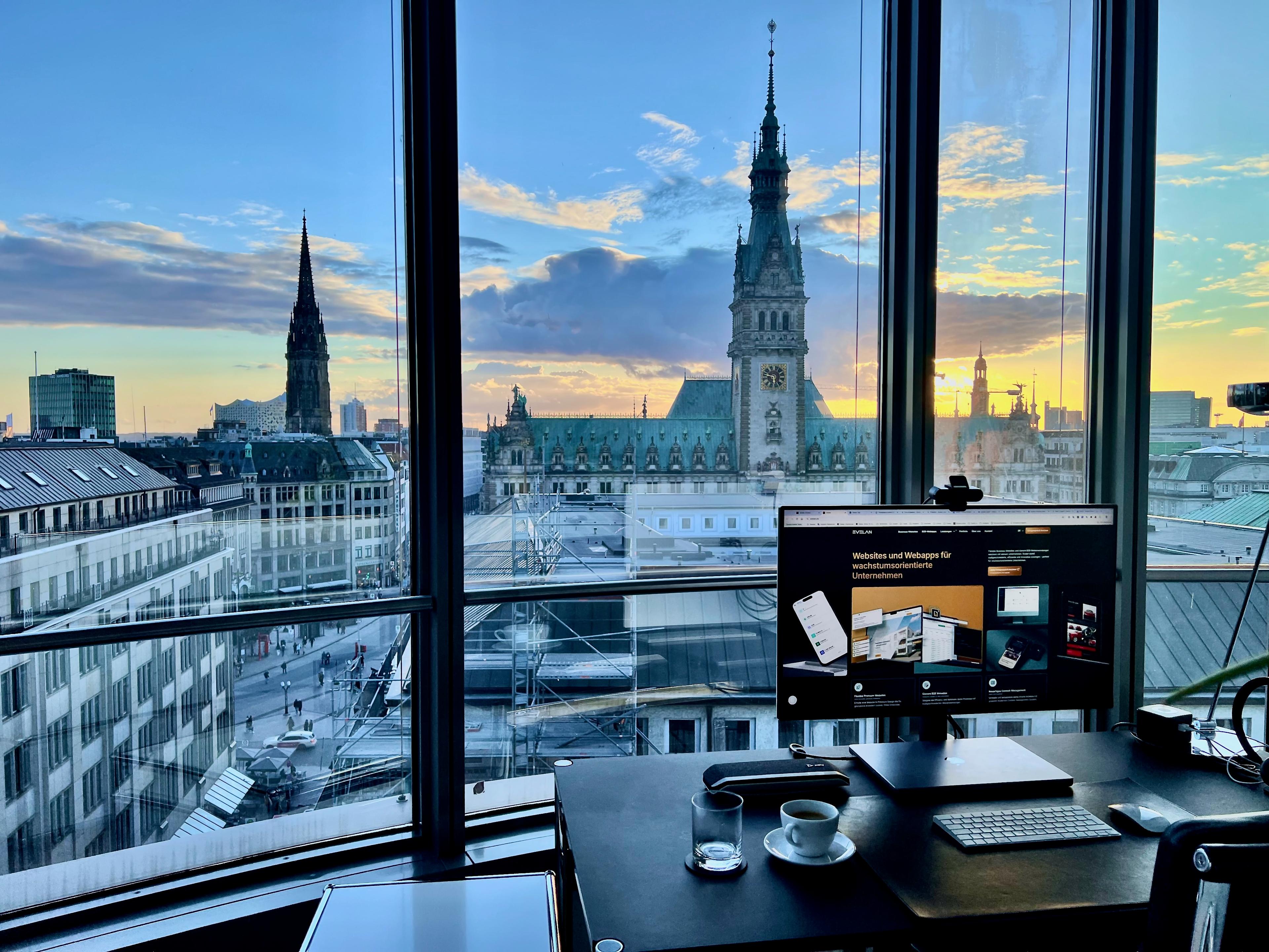 Office desk setup with a computer monitor and a view of a cityscape at sunset through large windows