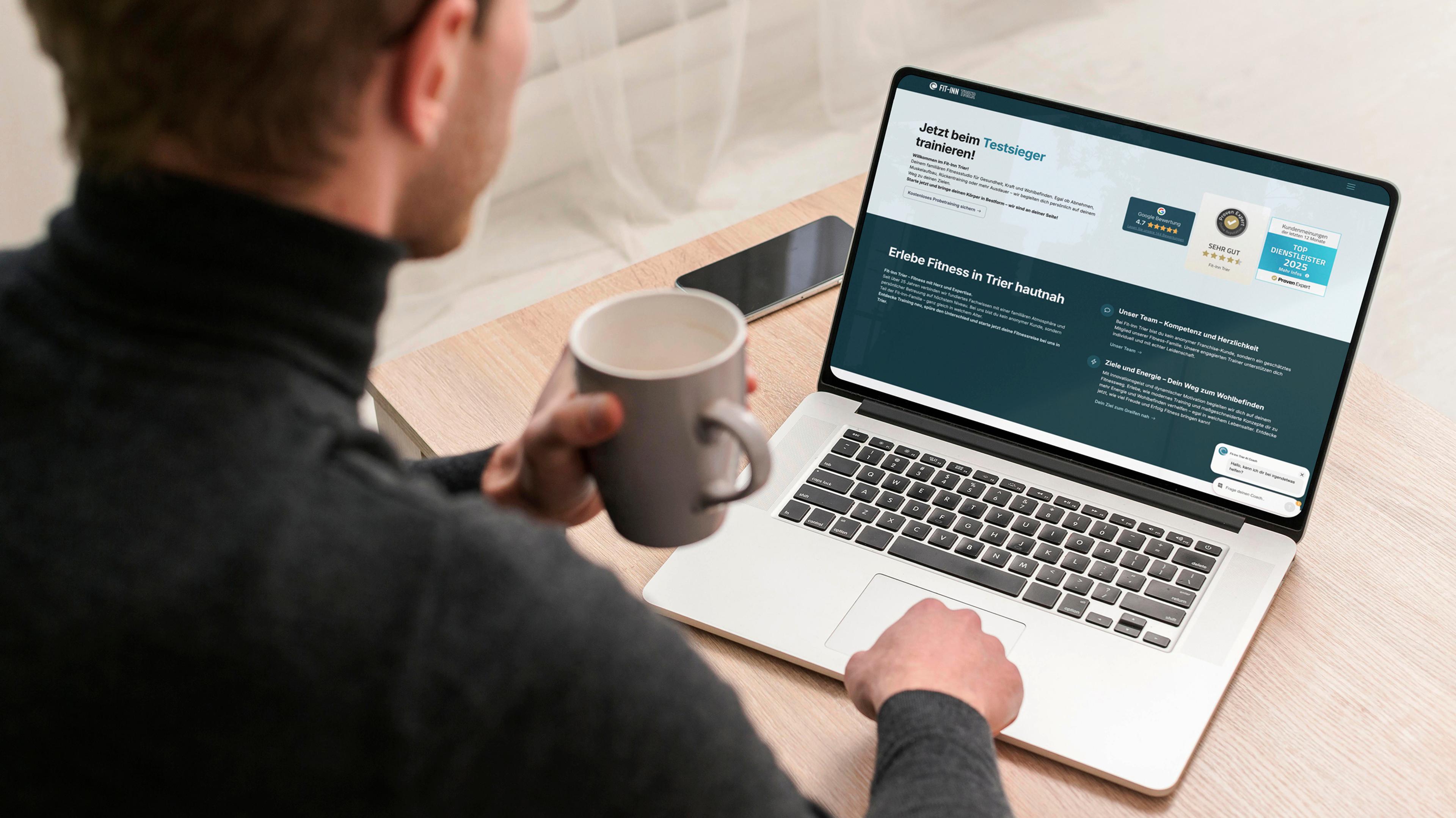 Mann in Rollkragenpullover mit grauer Kaffeetasse vor Laptop, Webseite sichtbar