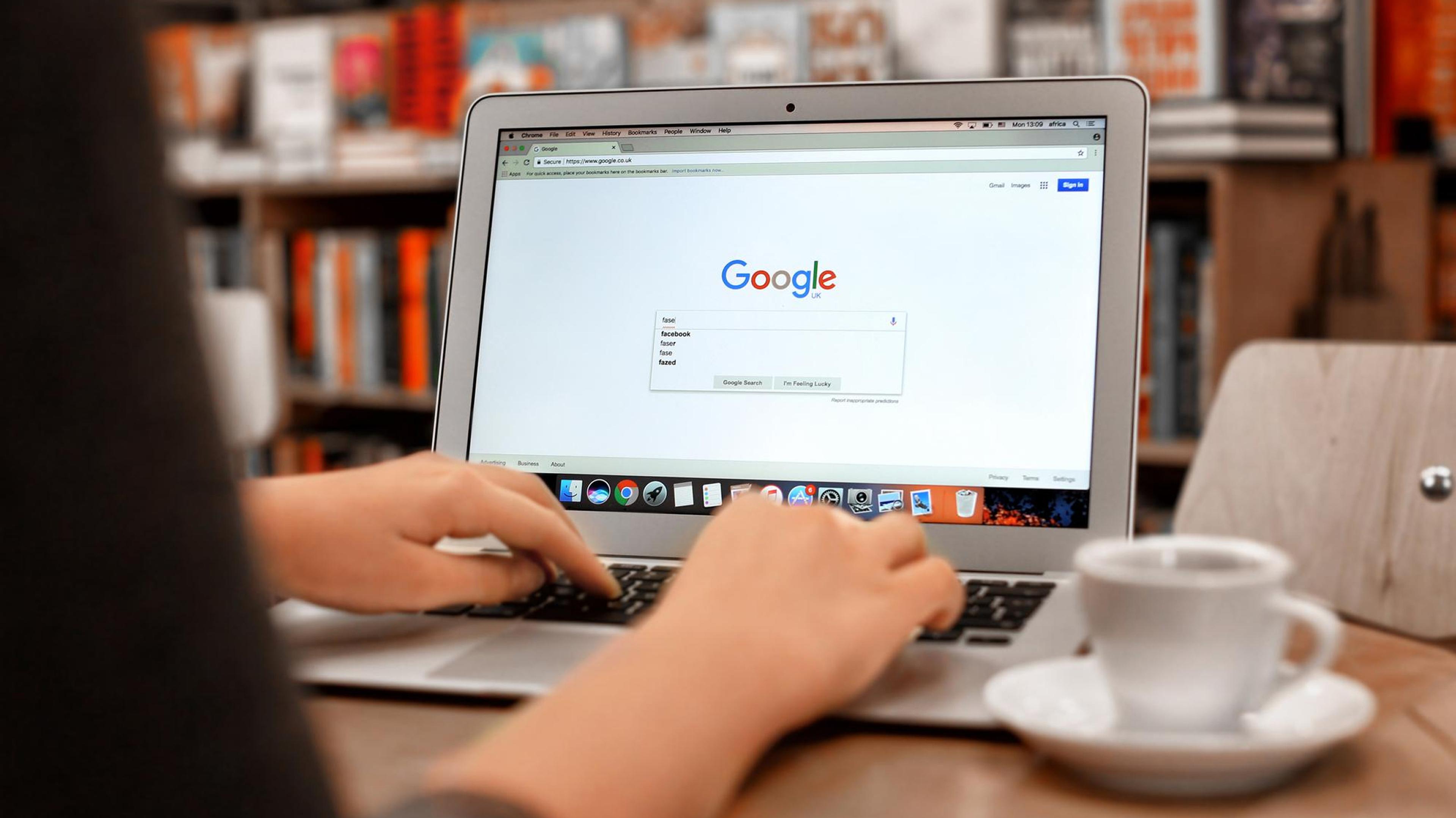 En person skriver på en bærbar PC i et bibliotek, med et Google-søk synlig, og en kaffekopp foran seg