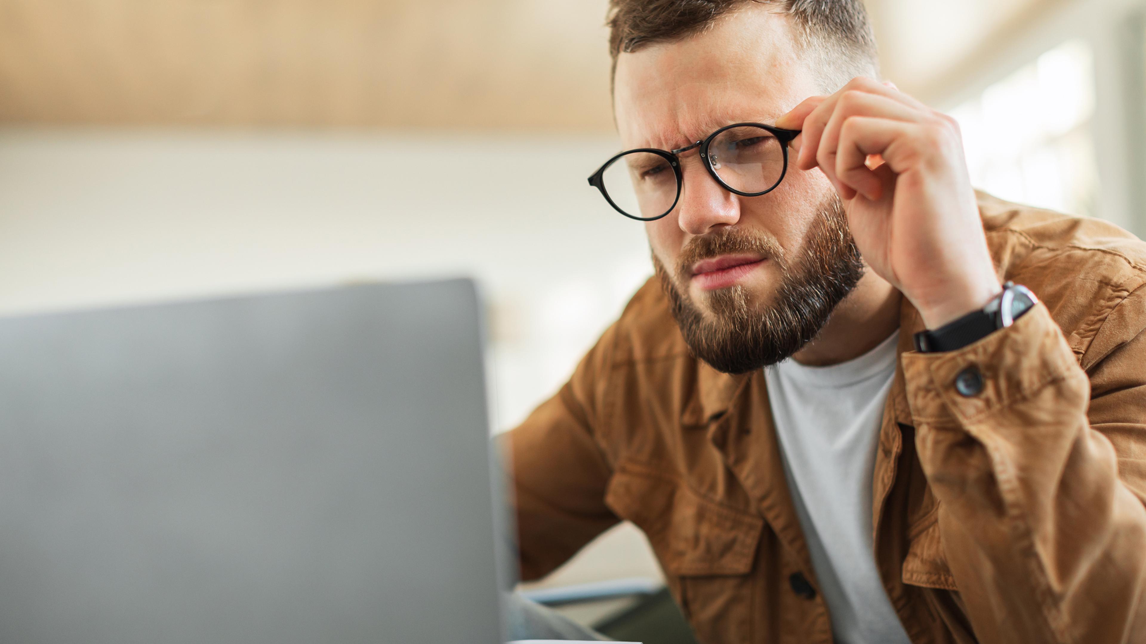 A man with glasses and a brown jacket looks pensively at a laptop