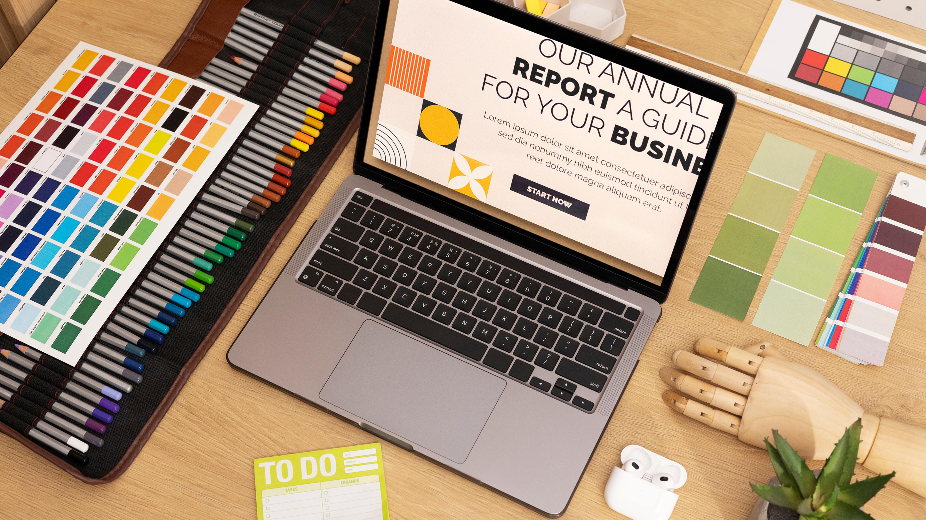 Laptop auf einem Holztisch umgeben von Farbmusterkarten, Stiften und einer hölzernen Hand.