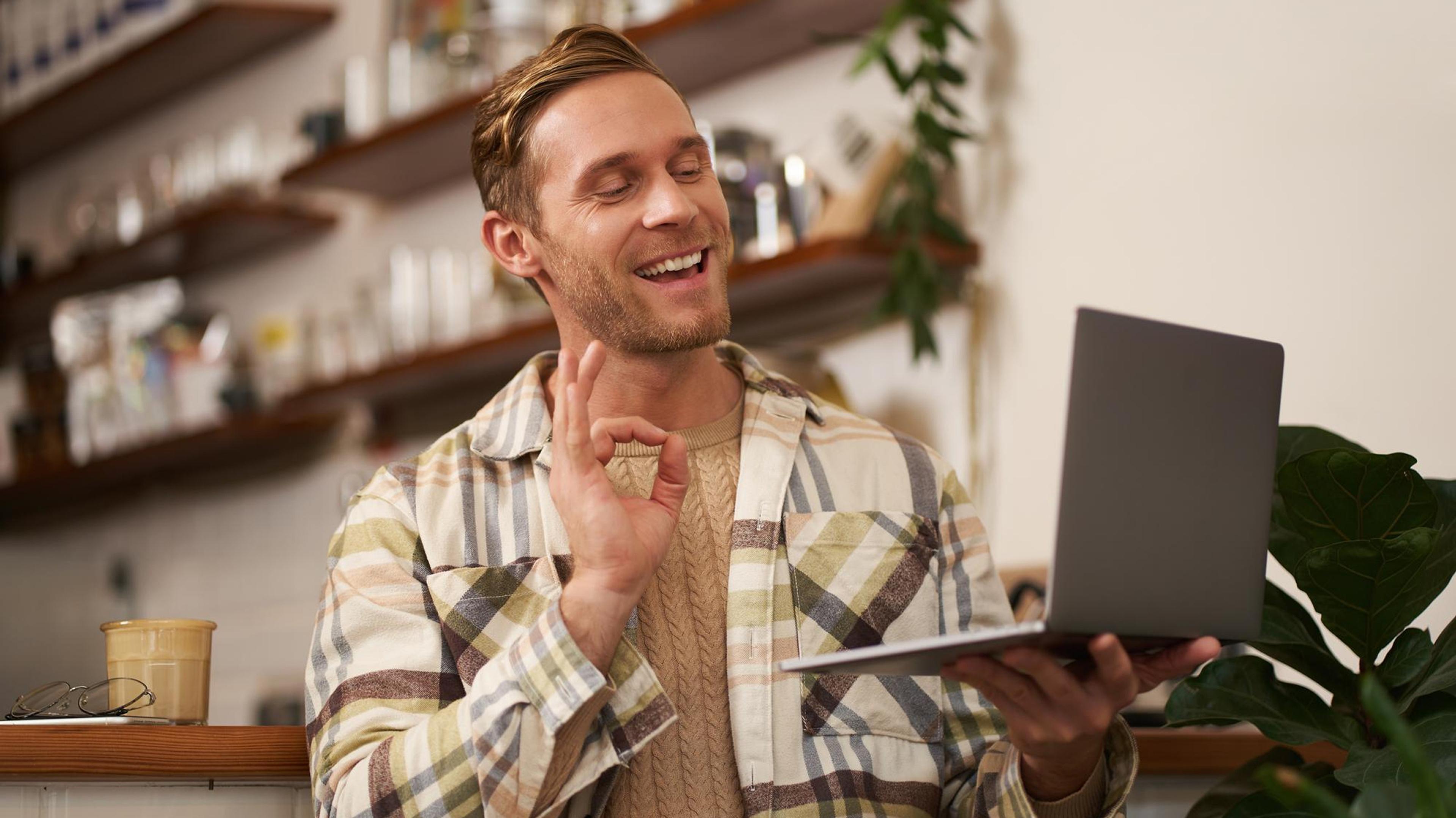 Mann im karierten Hemd mit Laptop, zeigt das Okay-Zeichen, im Café neben Kaffee, Pflanzen im Hintergrund.