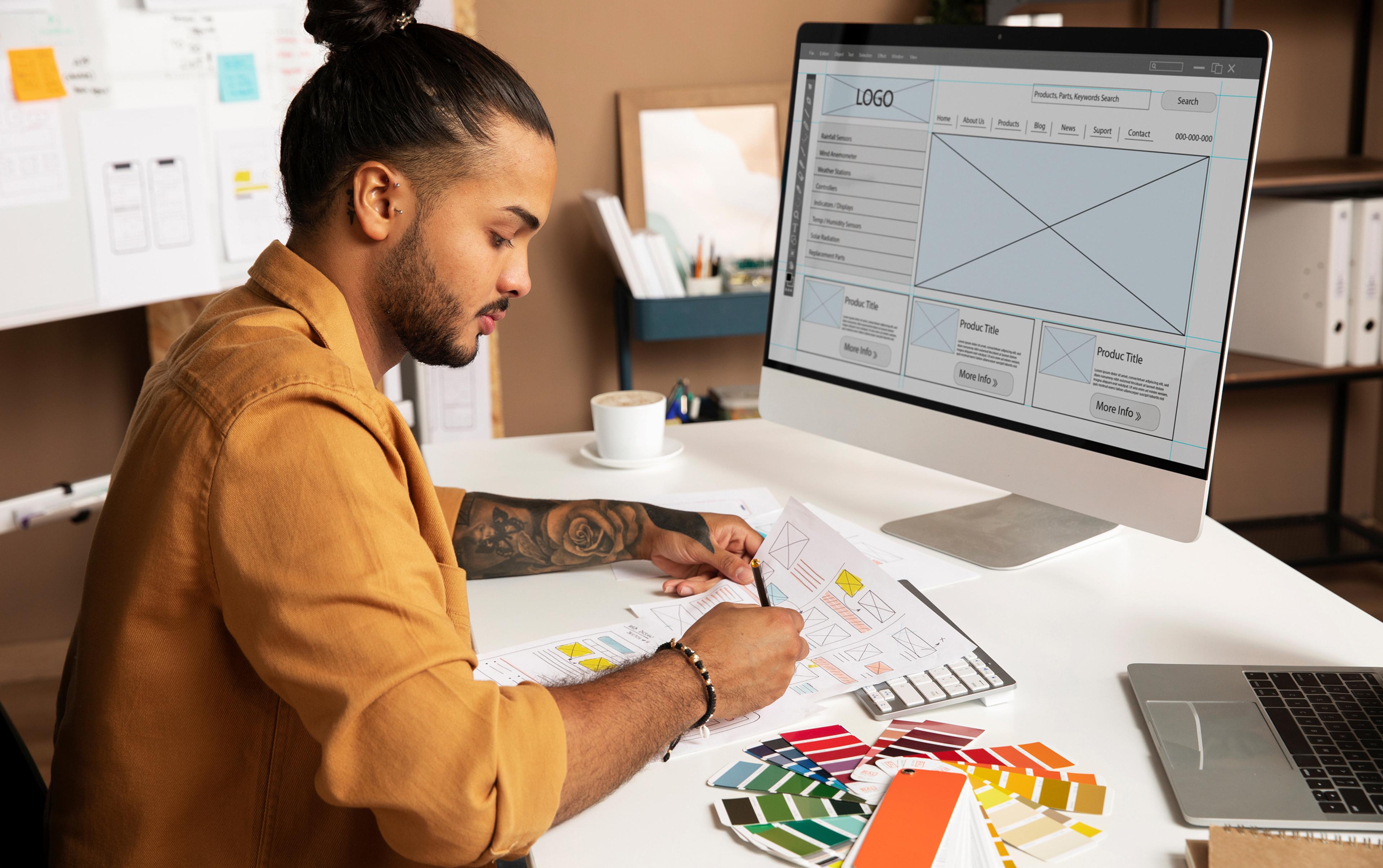 Mann in braunem Hemd arbeitet an Webdesign-Entwürfen, mit Farbpaletten und Computer auf dem Tisch.