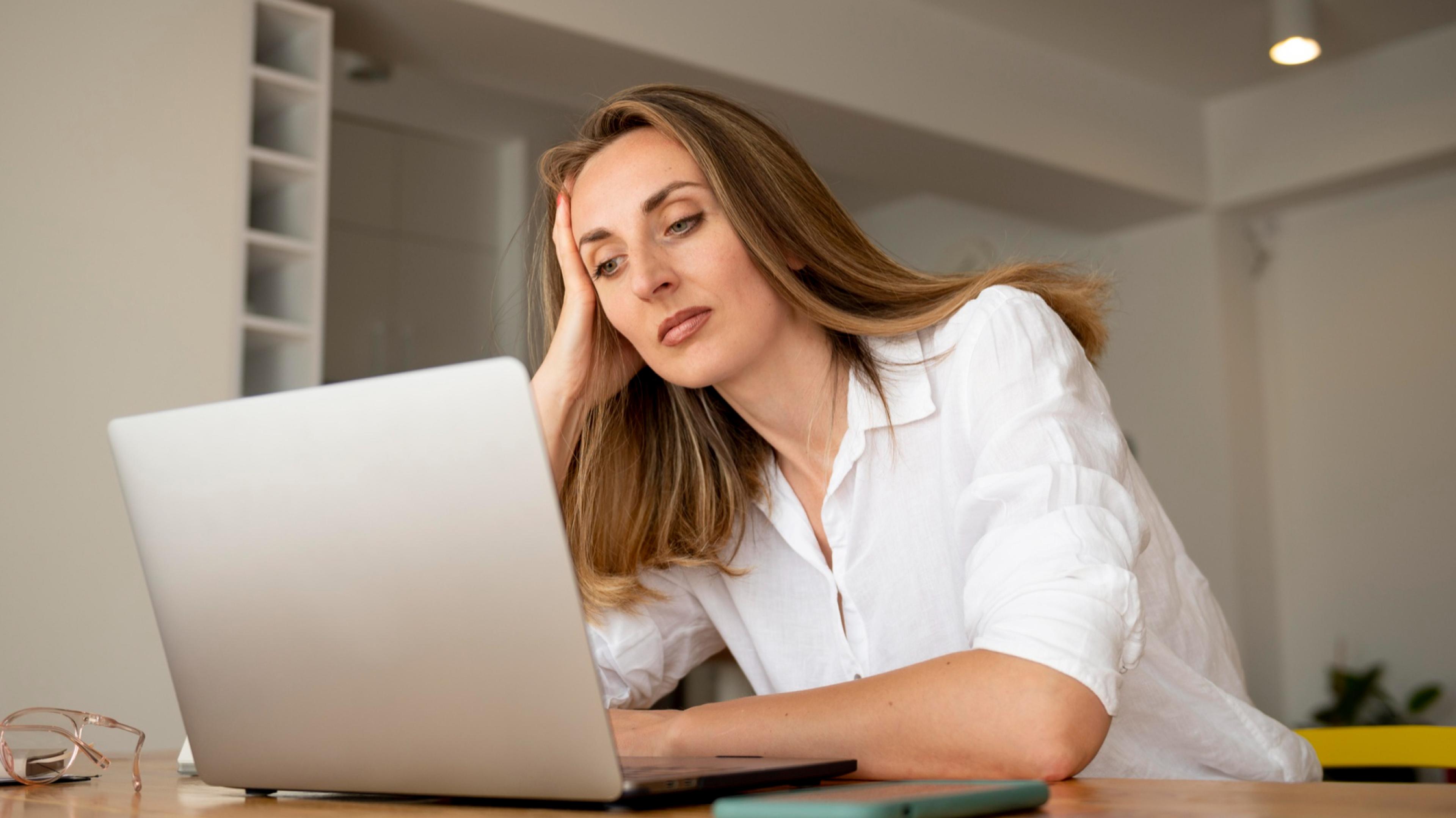 Eine Frau sitzt an einem Holztisch und schaut auf einen geöffneten silbernen Laptop, trägt ein weißes Hemd und stützt den Kopf mit der Hand; auf dem Tisch liegen eine Brille mit durchsichtigem Gestell und ein grünes Smartphone.