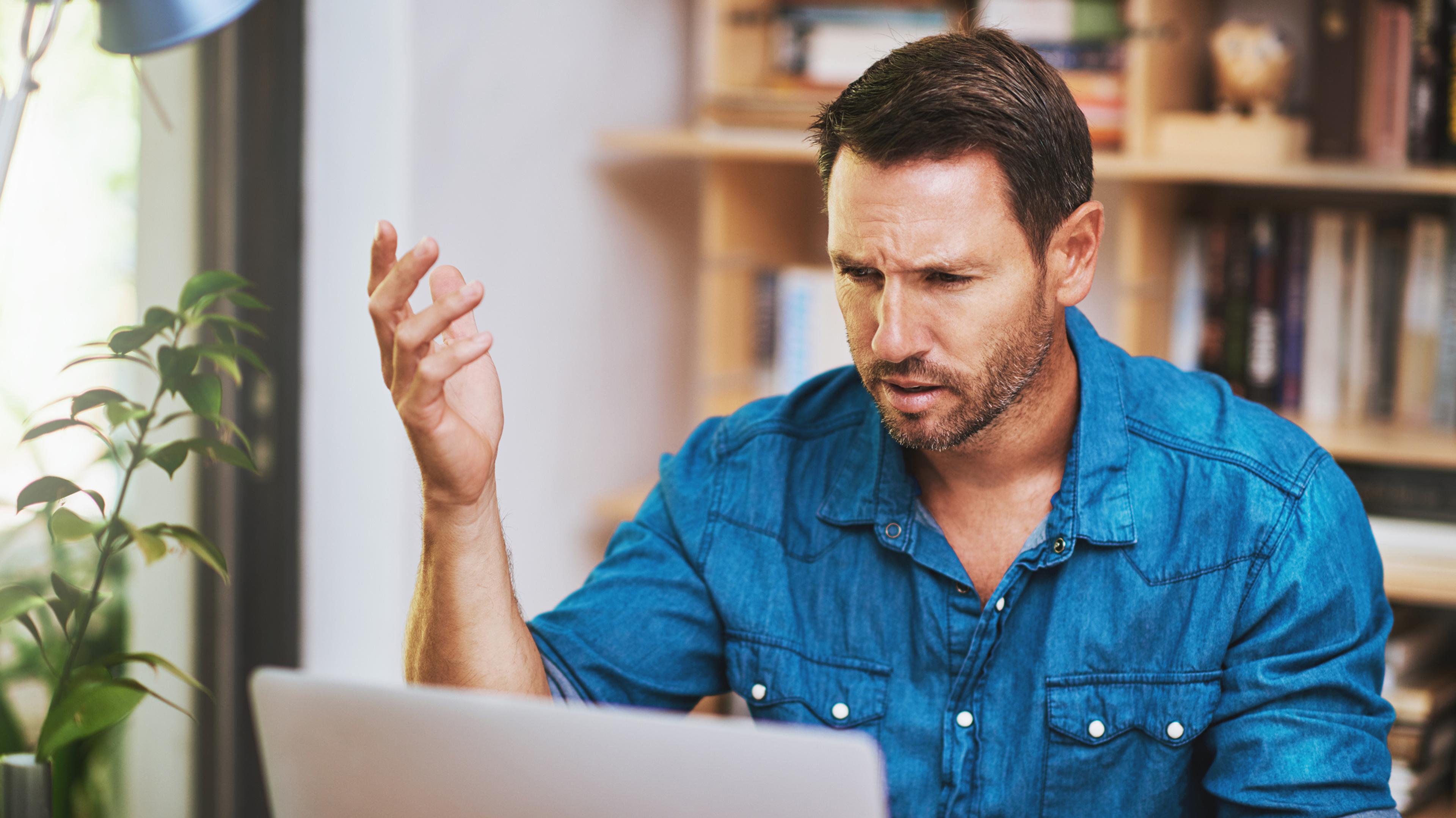 Mann in blauem Hemd sitzt vor einem Laptop, wirkt nachdenklich, Bücherregal im Hintergrund.