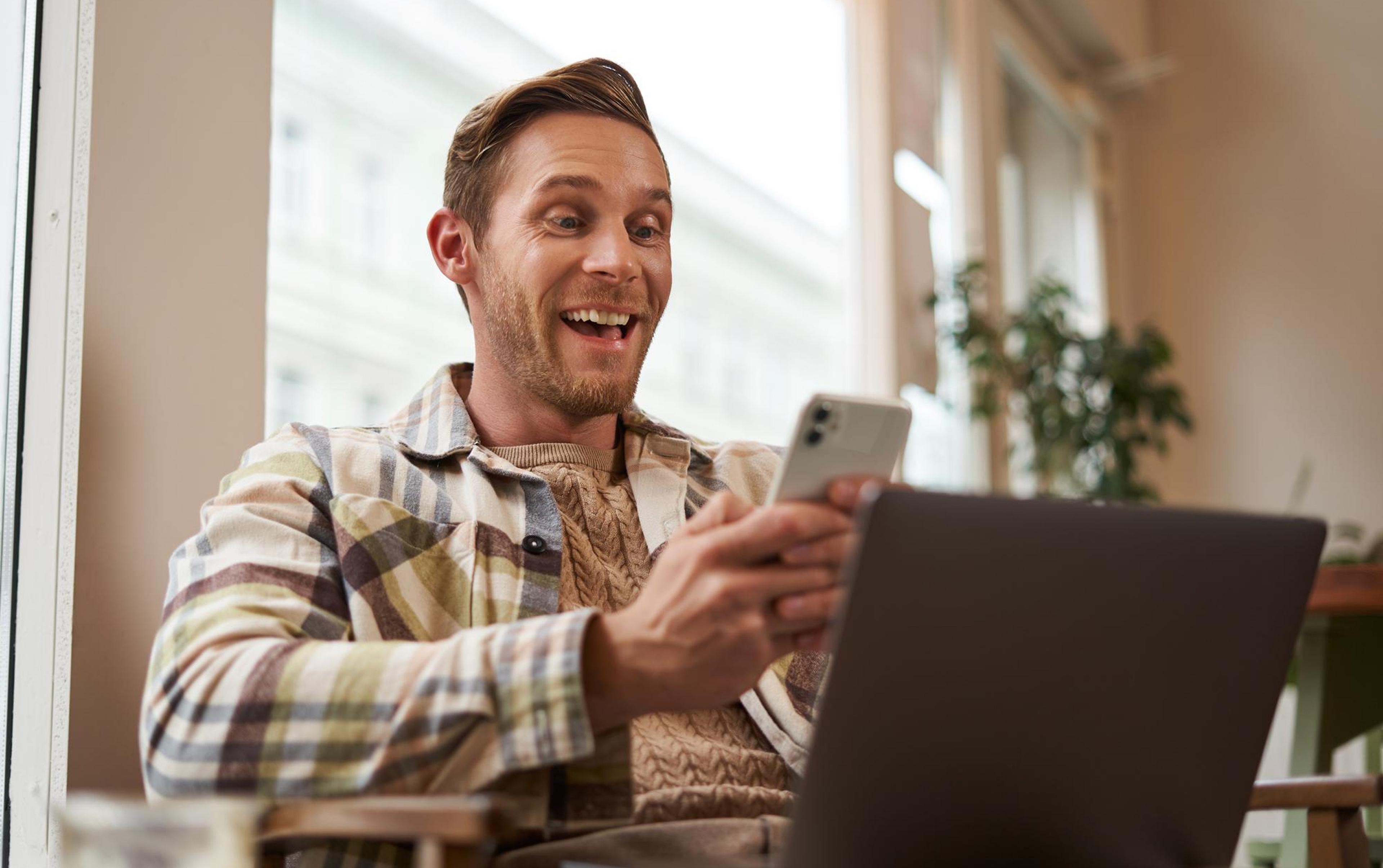 Mann mit kariertem Hemd sitzt lächelnd im Café, hält ein Smartphone und Laptop.