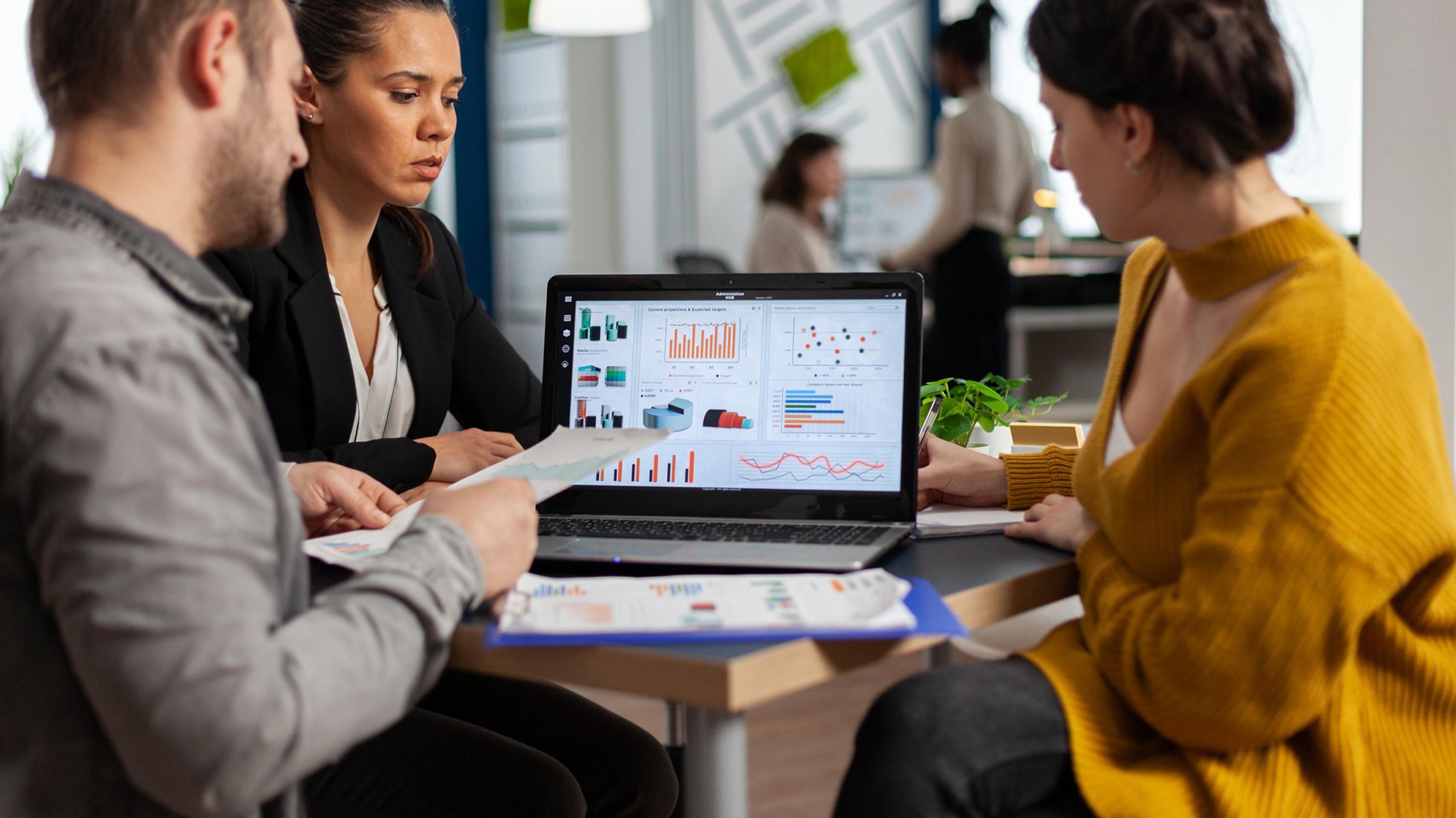 Drei Personen im Büro analysieren Diagramme auf einem Laptop. Hintergrund unscharf.