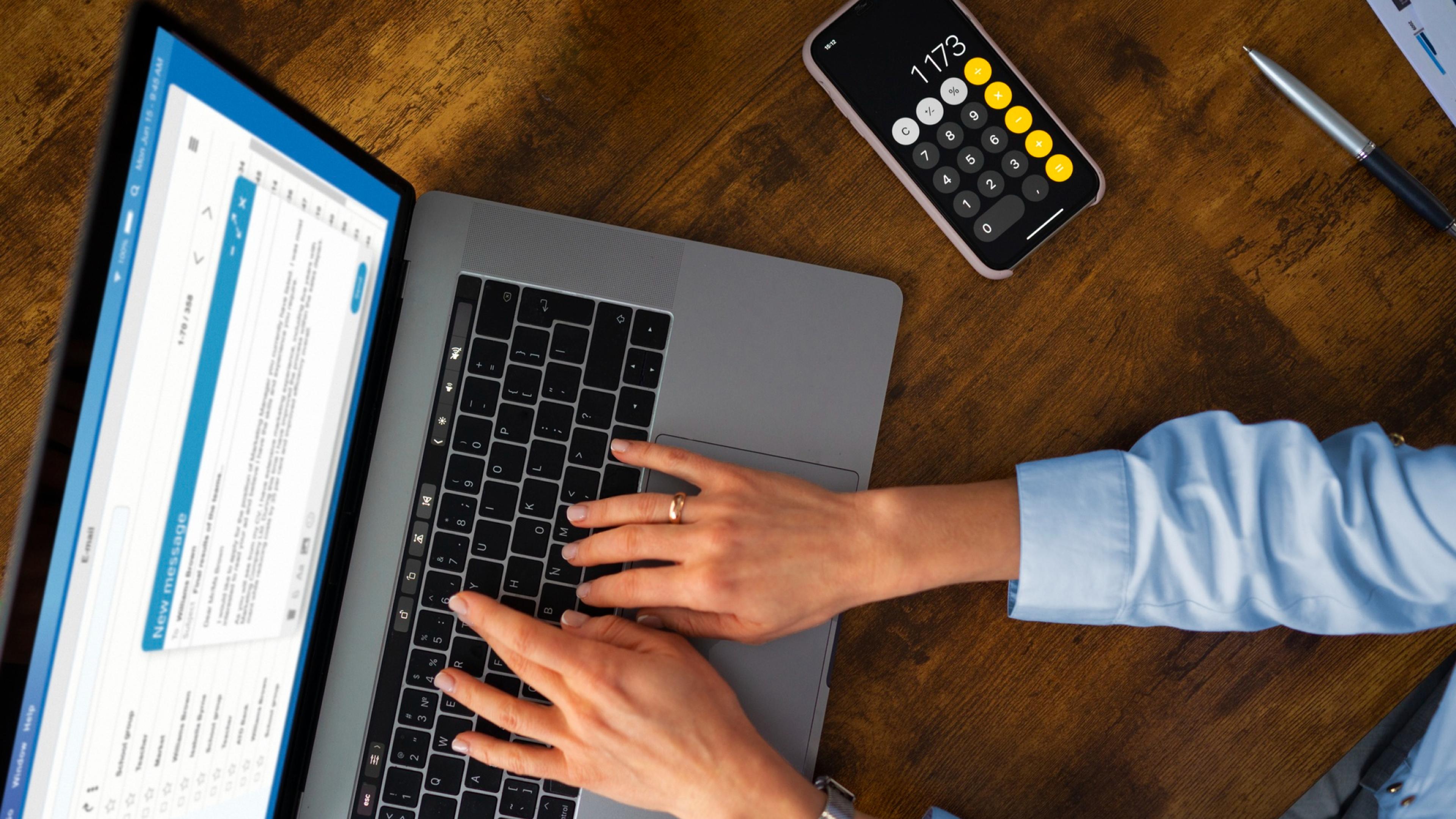 Hände tippen auf eine Laptop-Tastatur auf einem hölzernen Schreibtisch, daneben ein Smartphone mit geöffneter Taschenrechner-App
