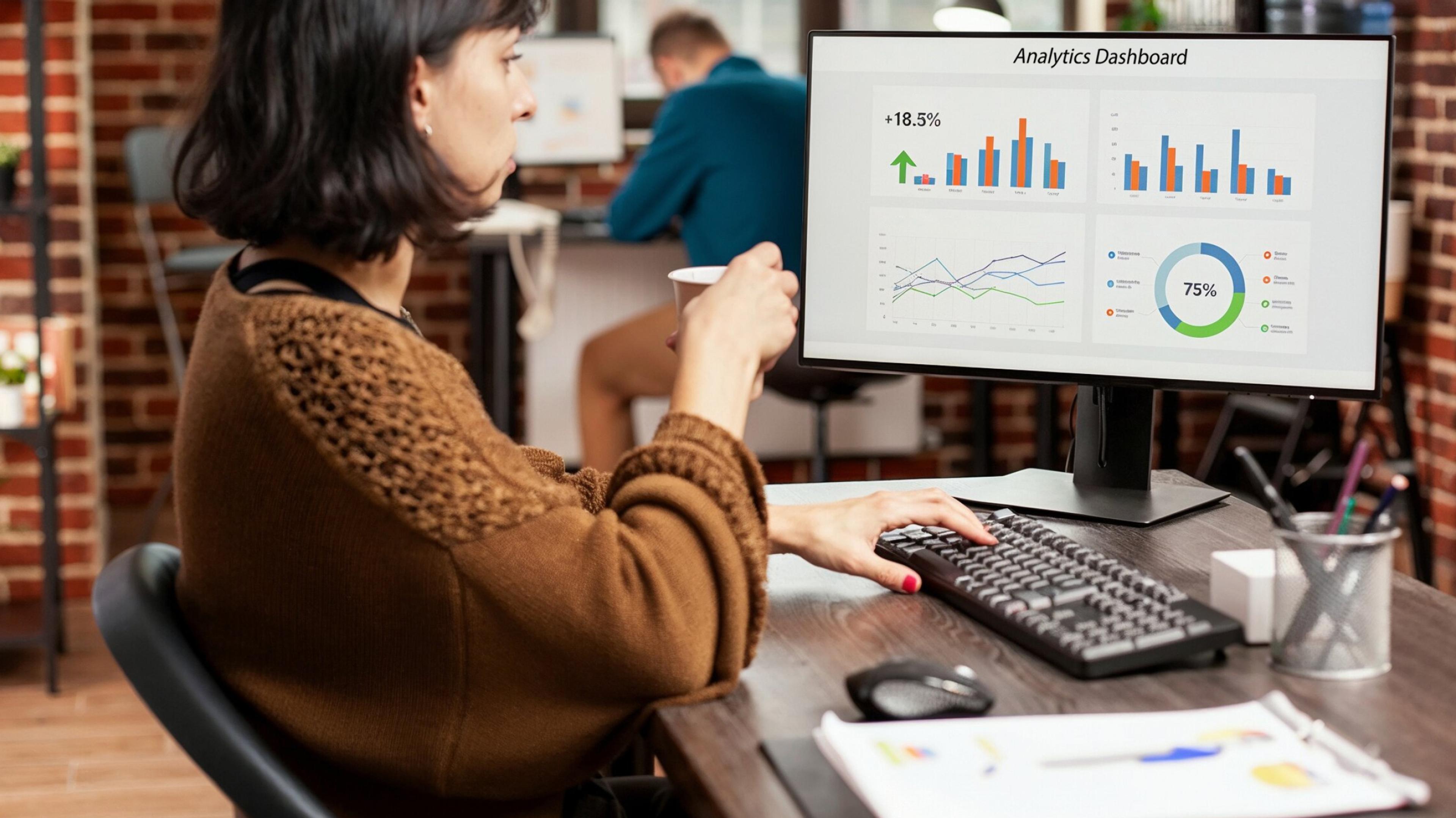Eine Person sitzt an einem Schreibtisch, hält eine Kaffeetasse und arbeitet mit der rechten Hand an einer Tastatur vor einem Monitor, der ein Analytics-Dashboard mit Balken-, Linien- und Kreisdiagramm anzeigt.
