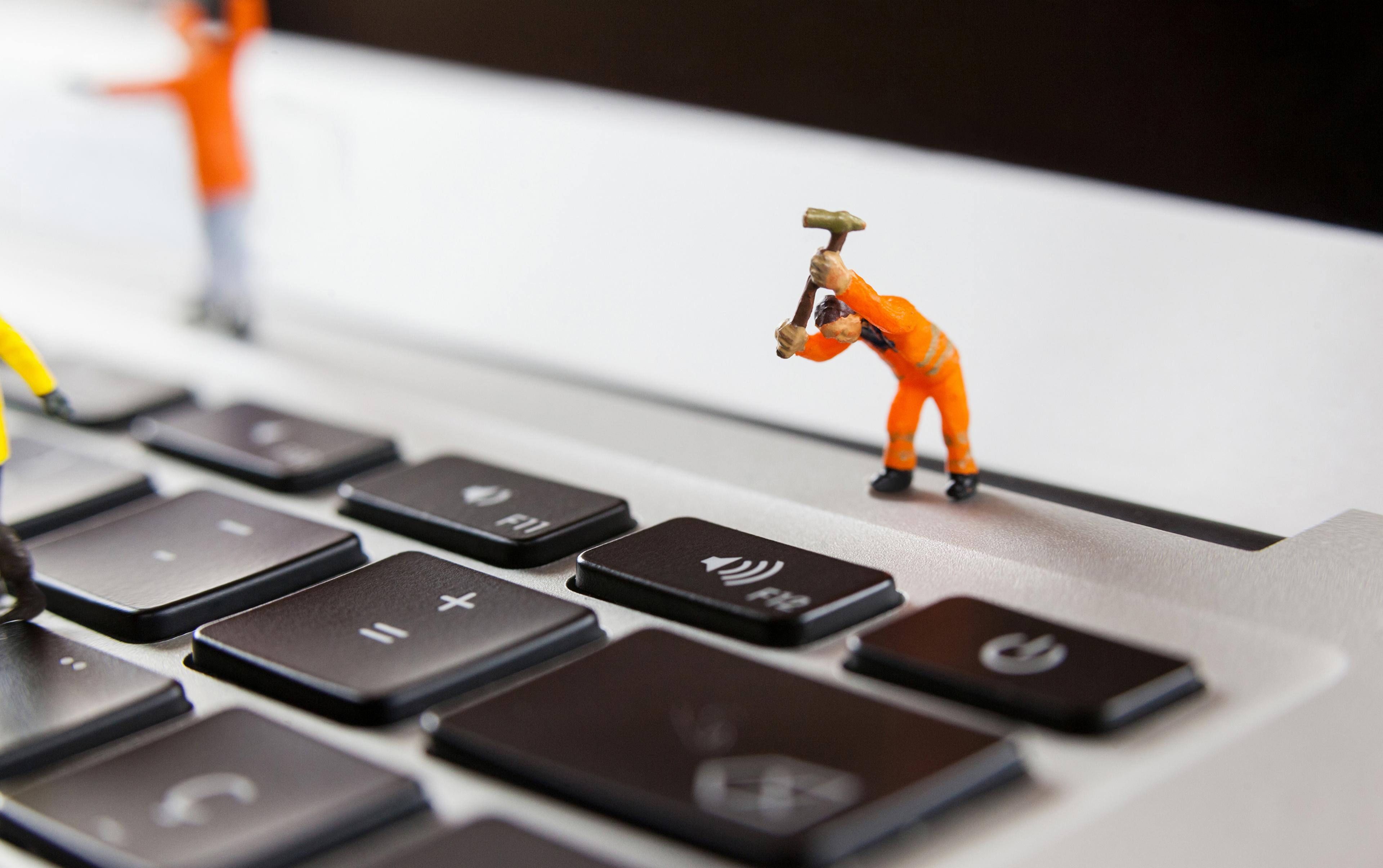 Ein Miniatur-Bauarbeiter in orangener Schutzkleidung hebt einen Hammer über einer Laptop-Taste