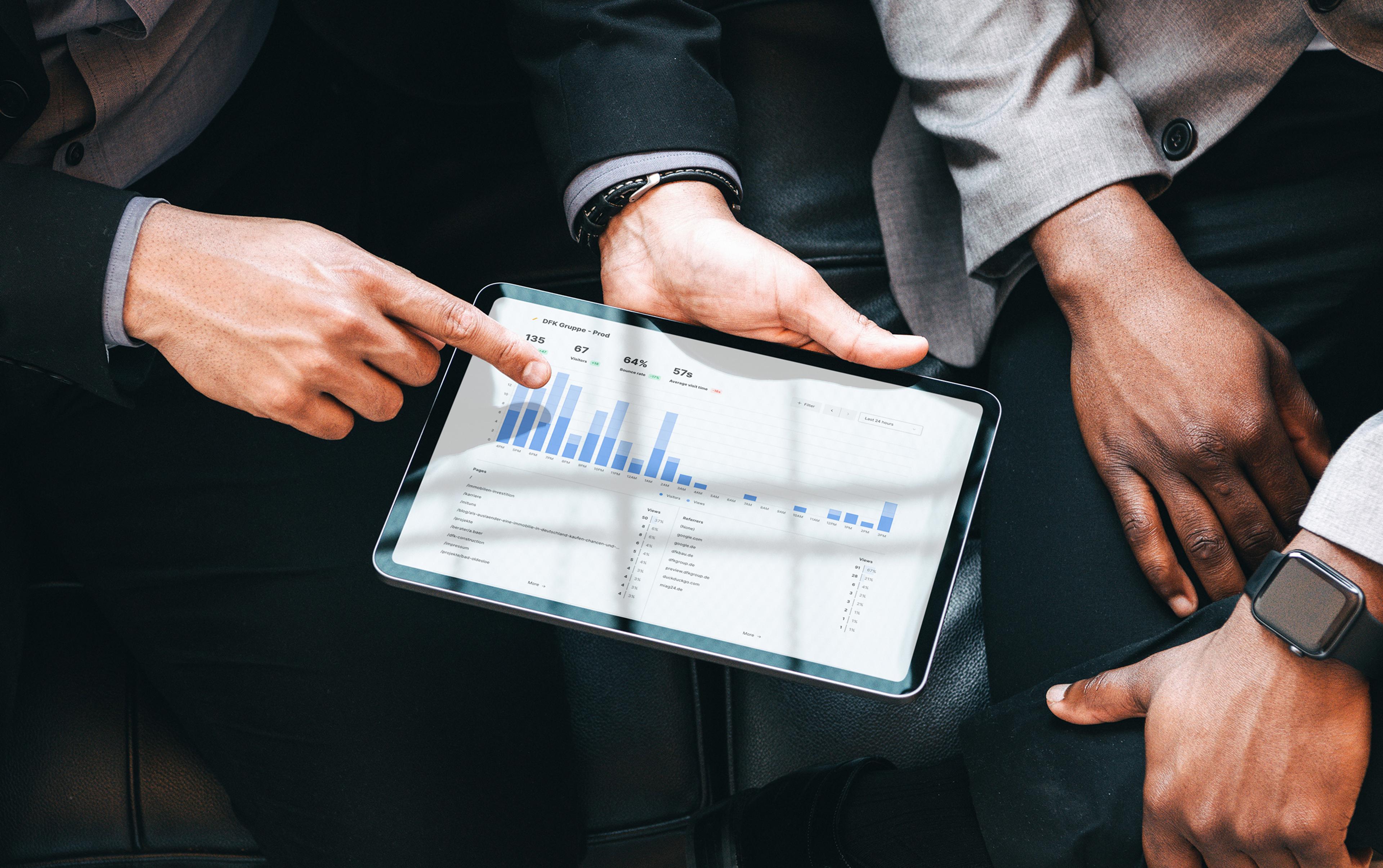 A tablet shows an analytics dashboard with blue bar charts and figures, while one person points to the display with their finger and a second hand holds the device.