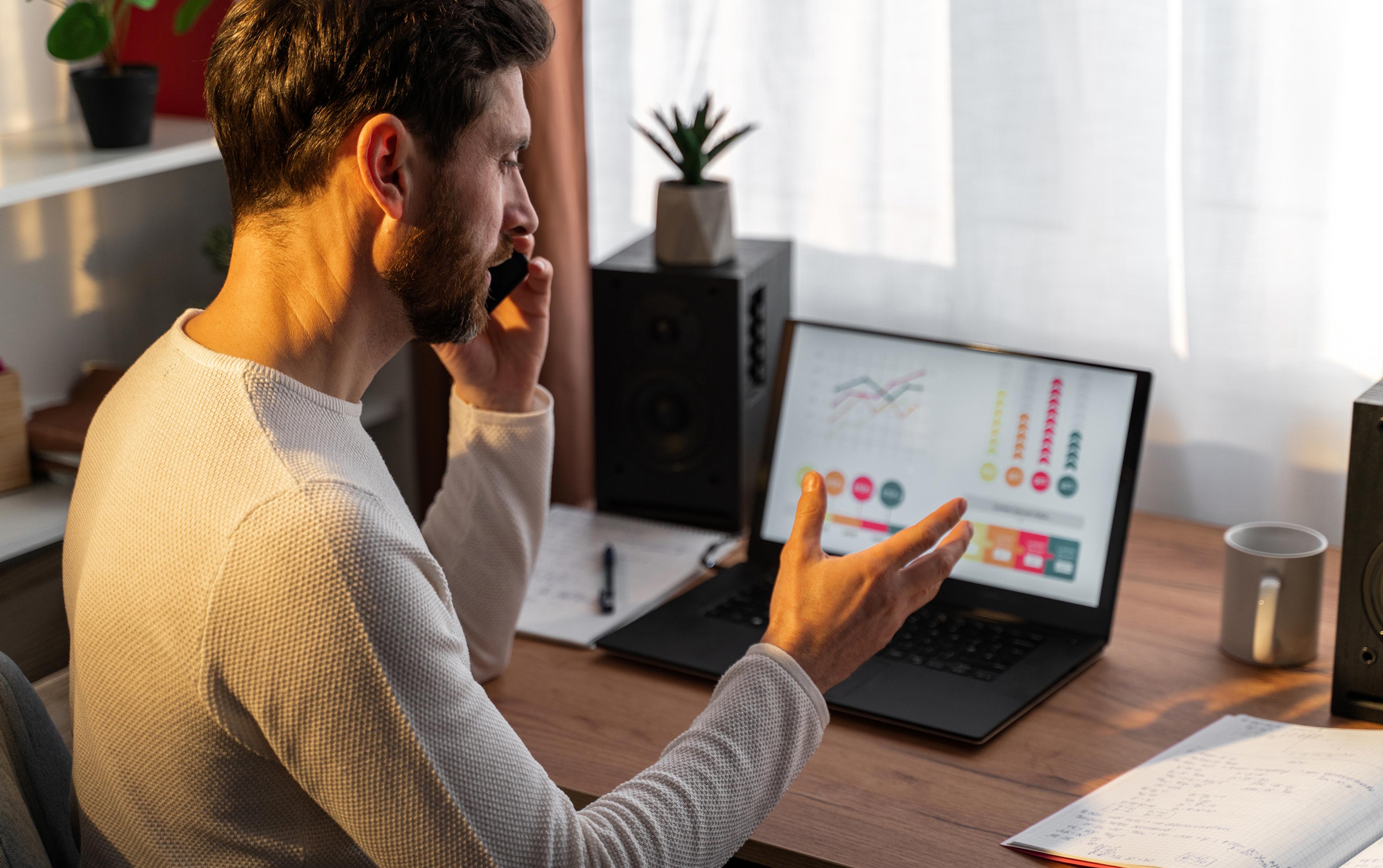 Man in home office on the phone, looking at laptop with diagrams, bright surroundings, potted plant in the background.