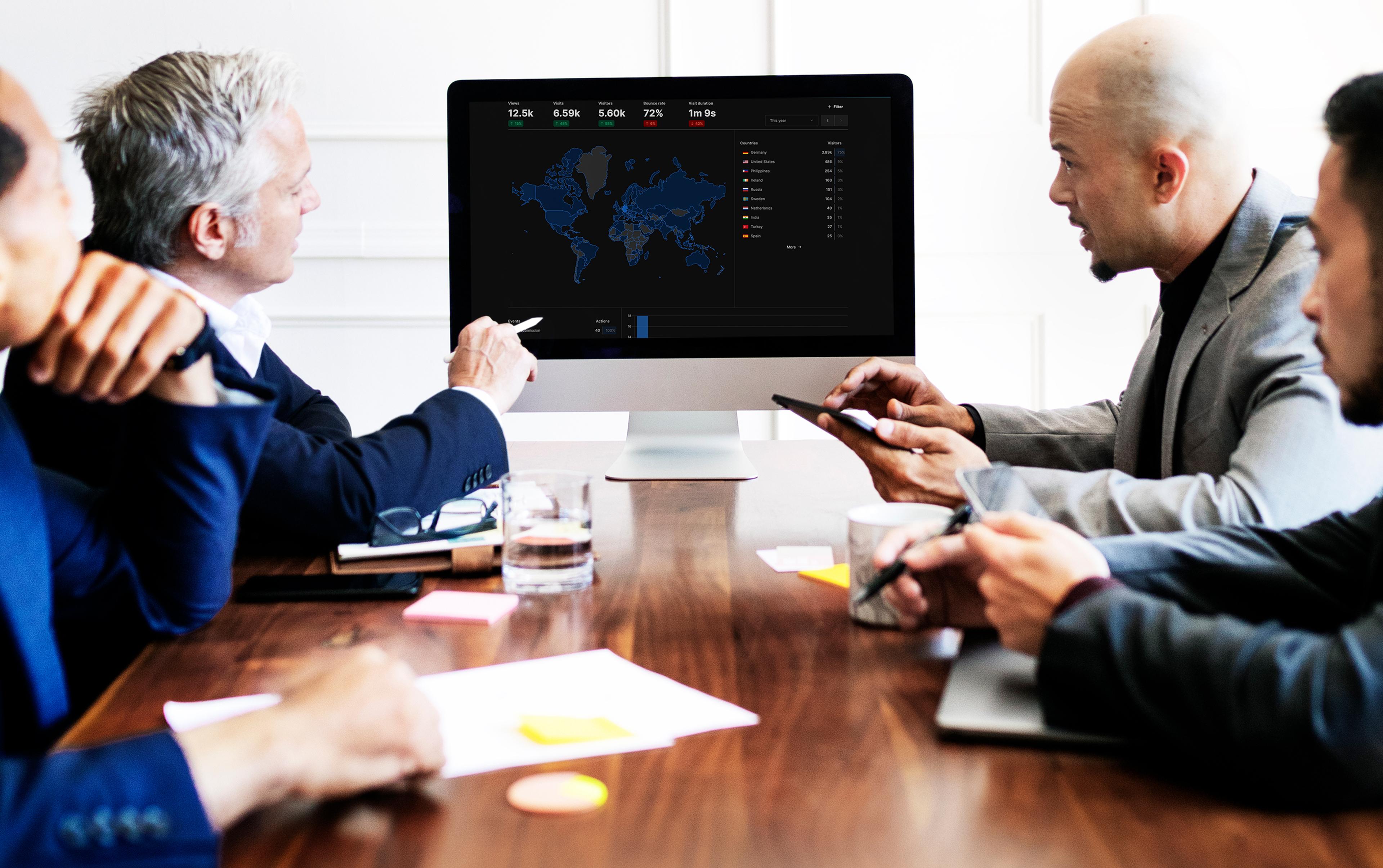 A team of four business people at a meeting in front of a large monitor showing a dark analytics interface with a world map and key figures.