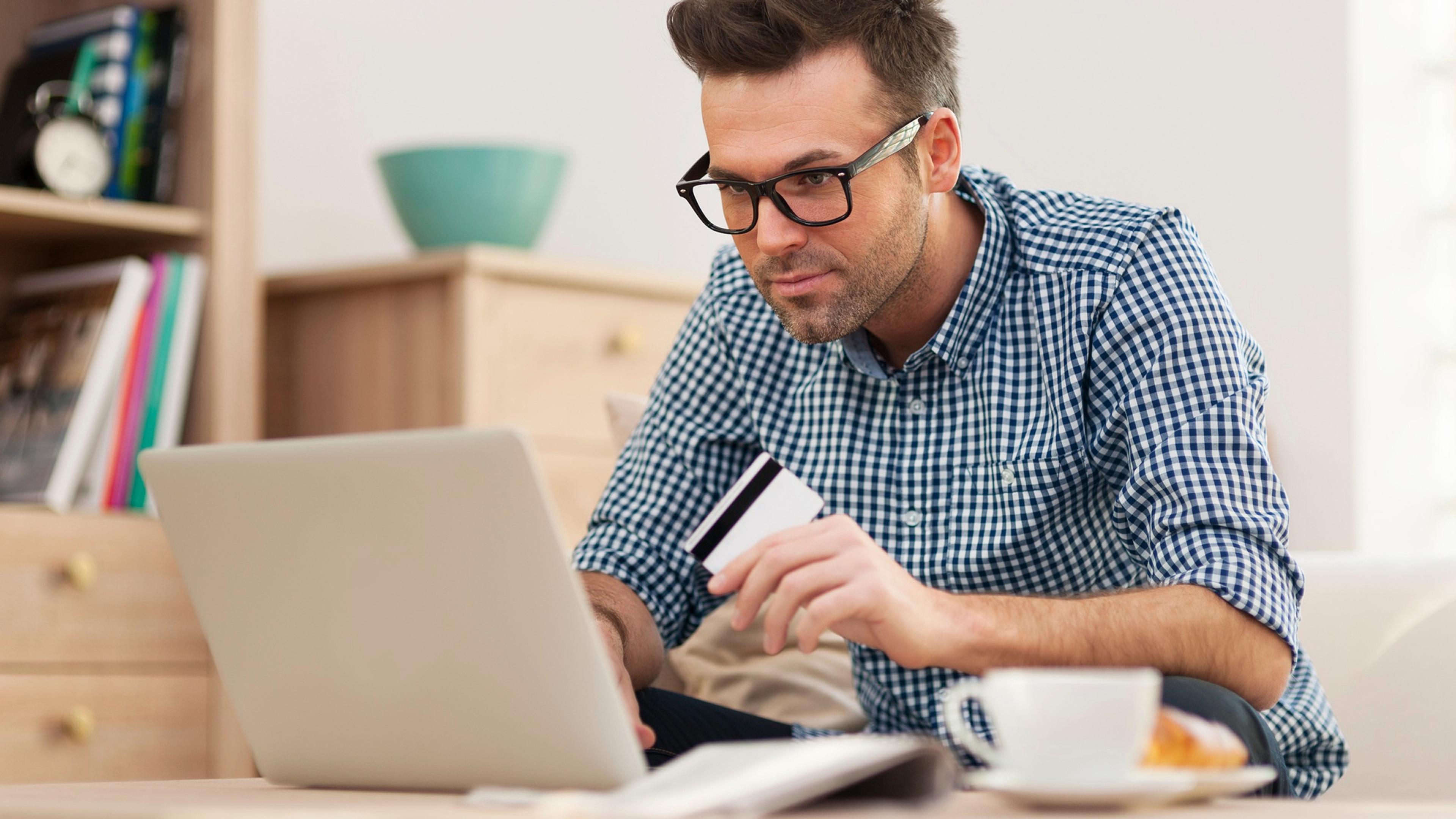 A man in a plaid shirt is holding a credit card and looking at a laptop in the living room.