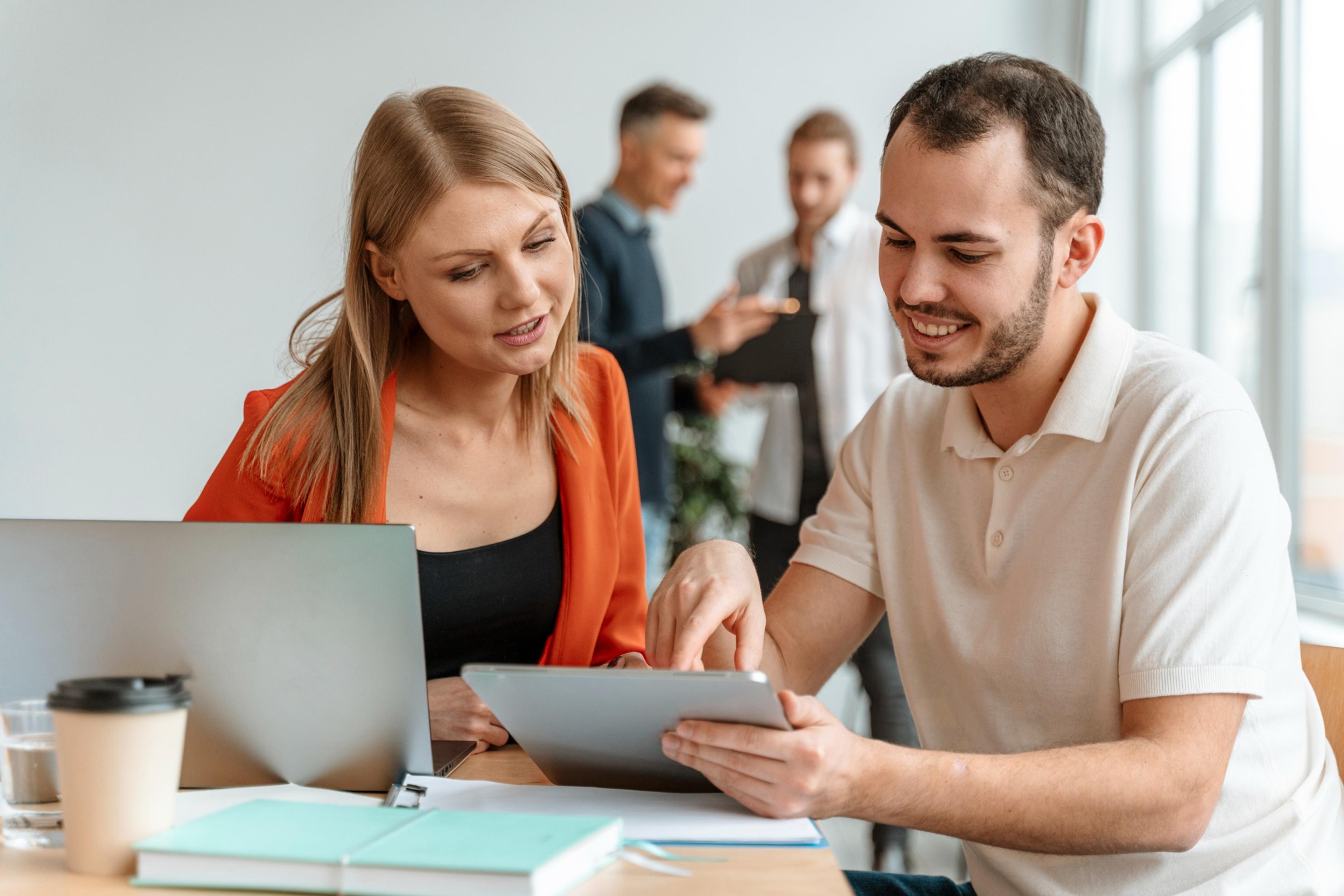 Zwei Personen diskutieren über ein Tablet, im Hintergrund weitere Personen in einem Büro.