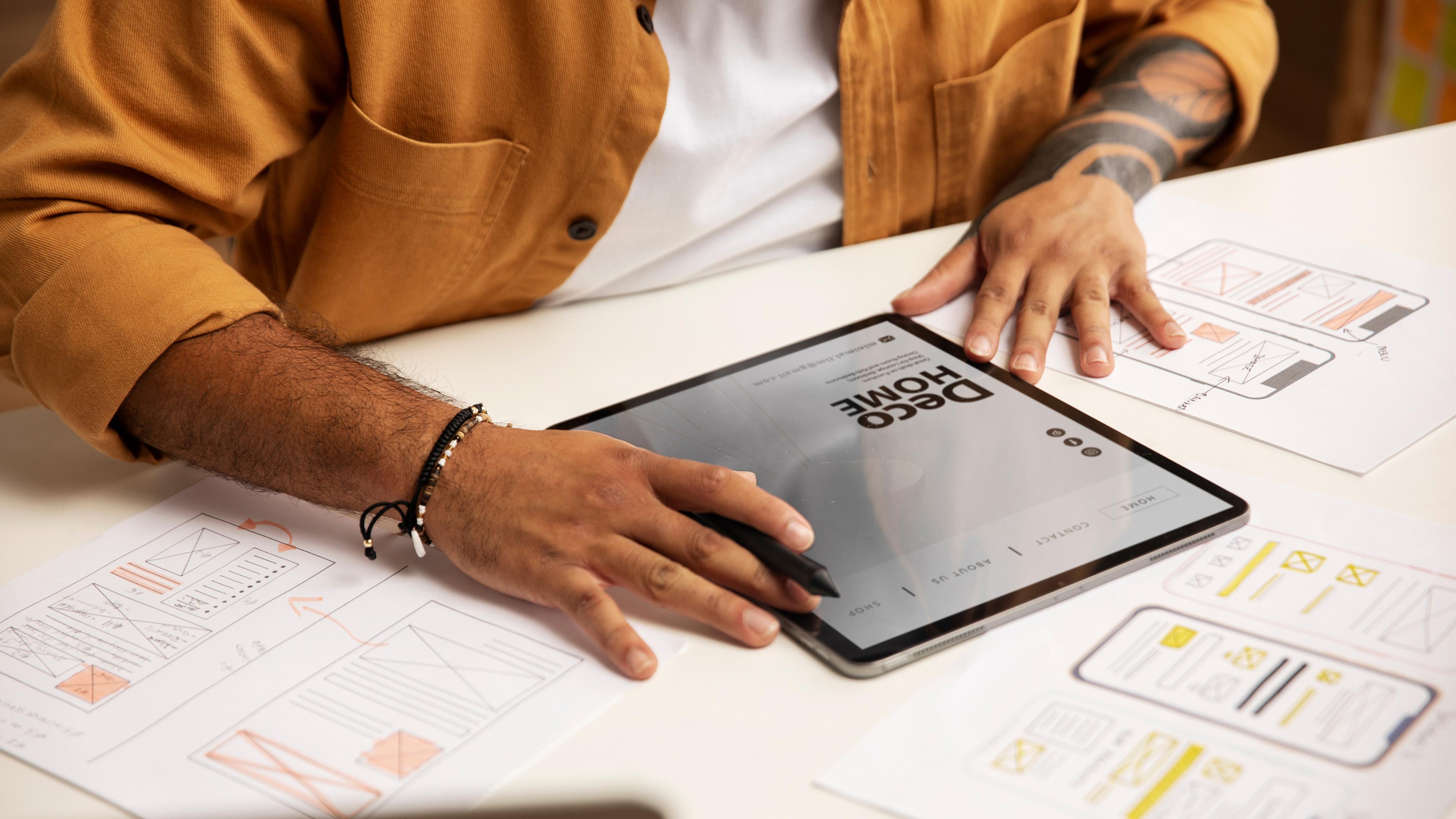 Mann in braunem Hemd benutzt ein Tablet für Prototypenentwurf mit Skizzen auf dem Tisch.