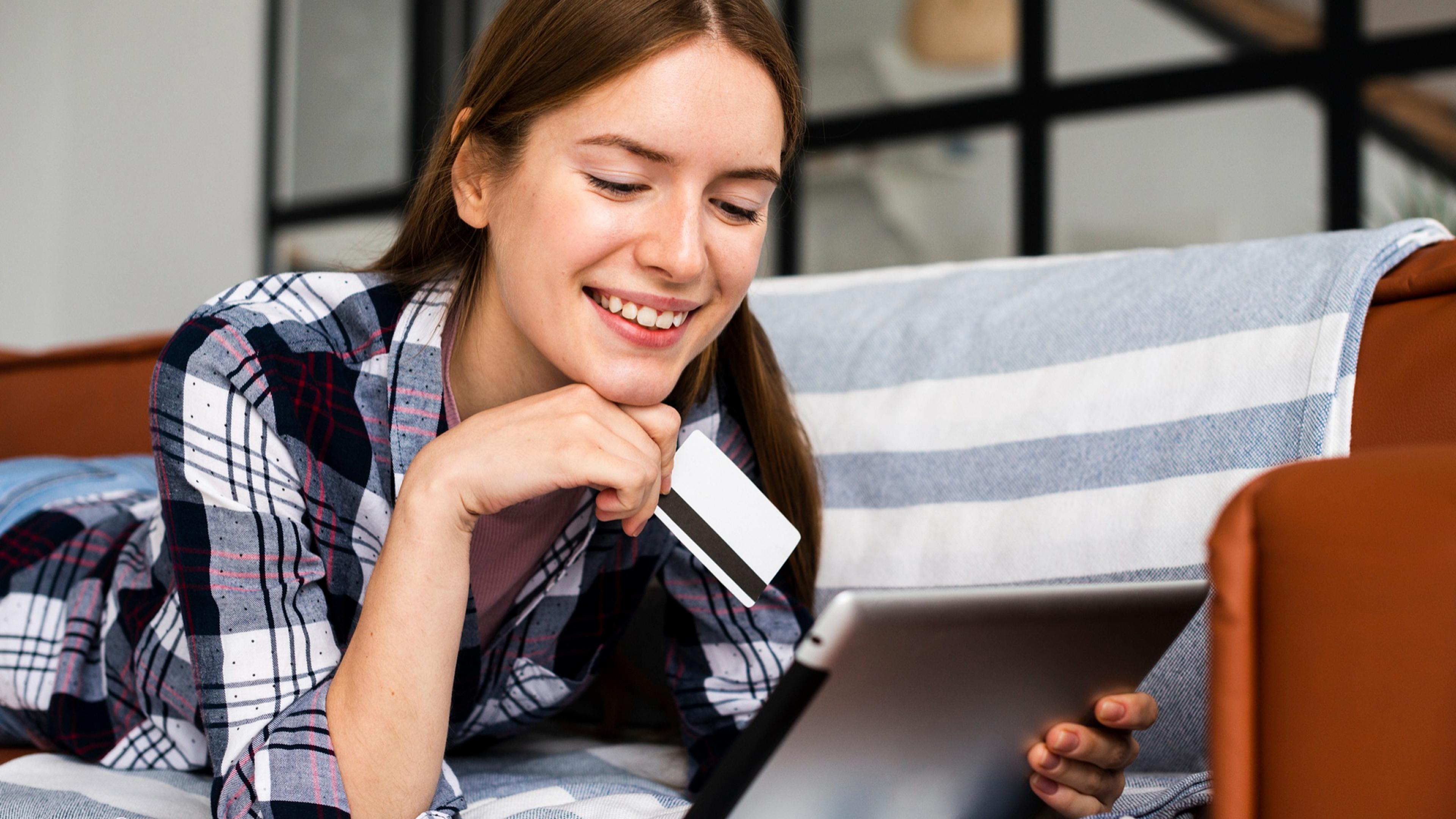 Frau in kariertem Hemd liegt auf Sofa, hält Kreditkarte und schaut auf Tablet.