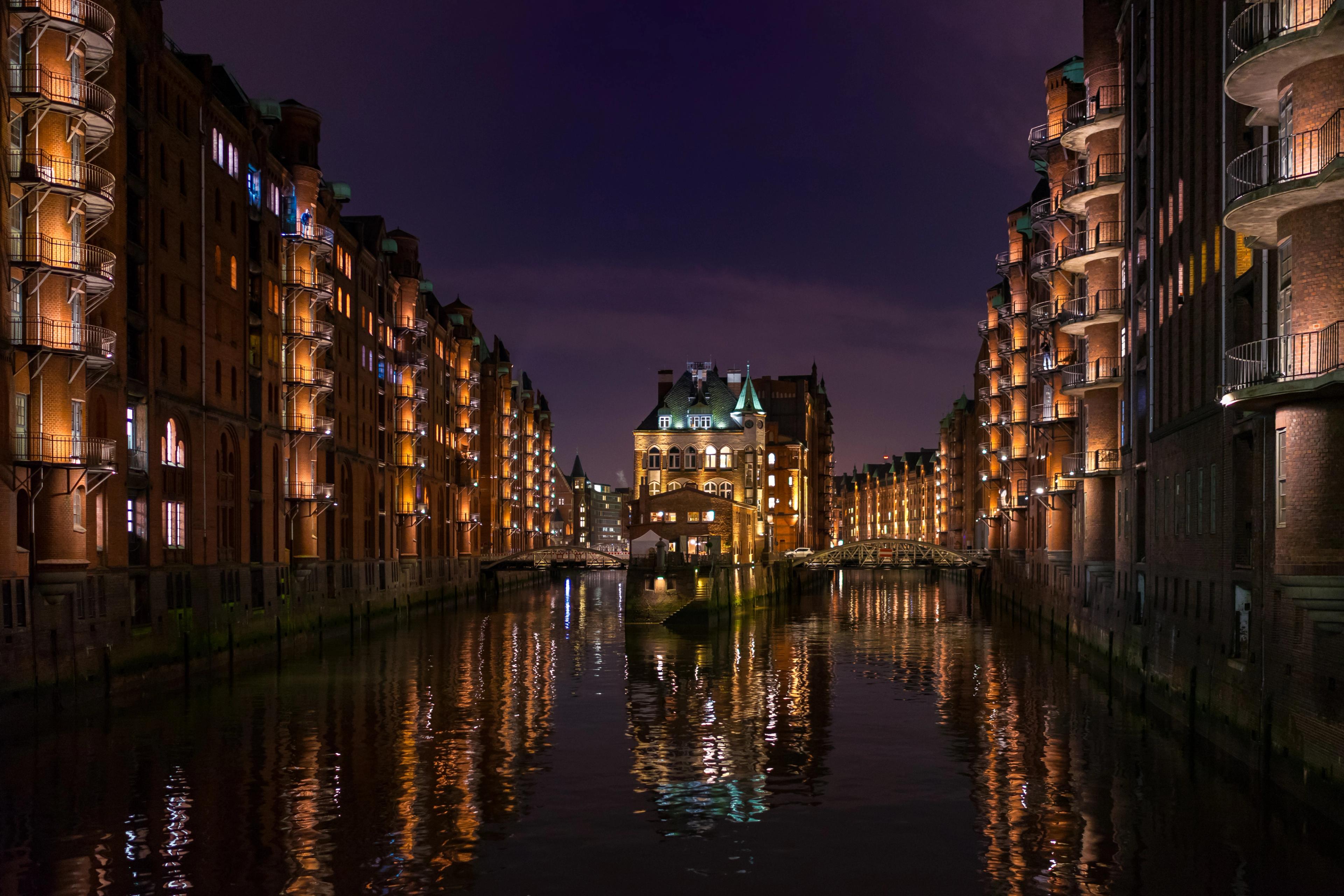 Ein nächtlicher Blick auf einen beleuchteten Kanal zwischen roten Backsteingebäuden, mit Brücken, Balkonen und spiegelnden Lichtern im Wasser.