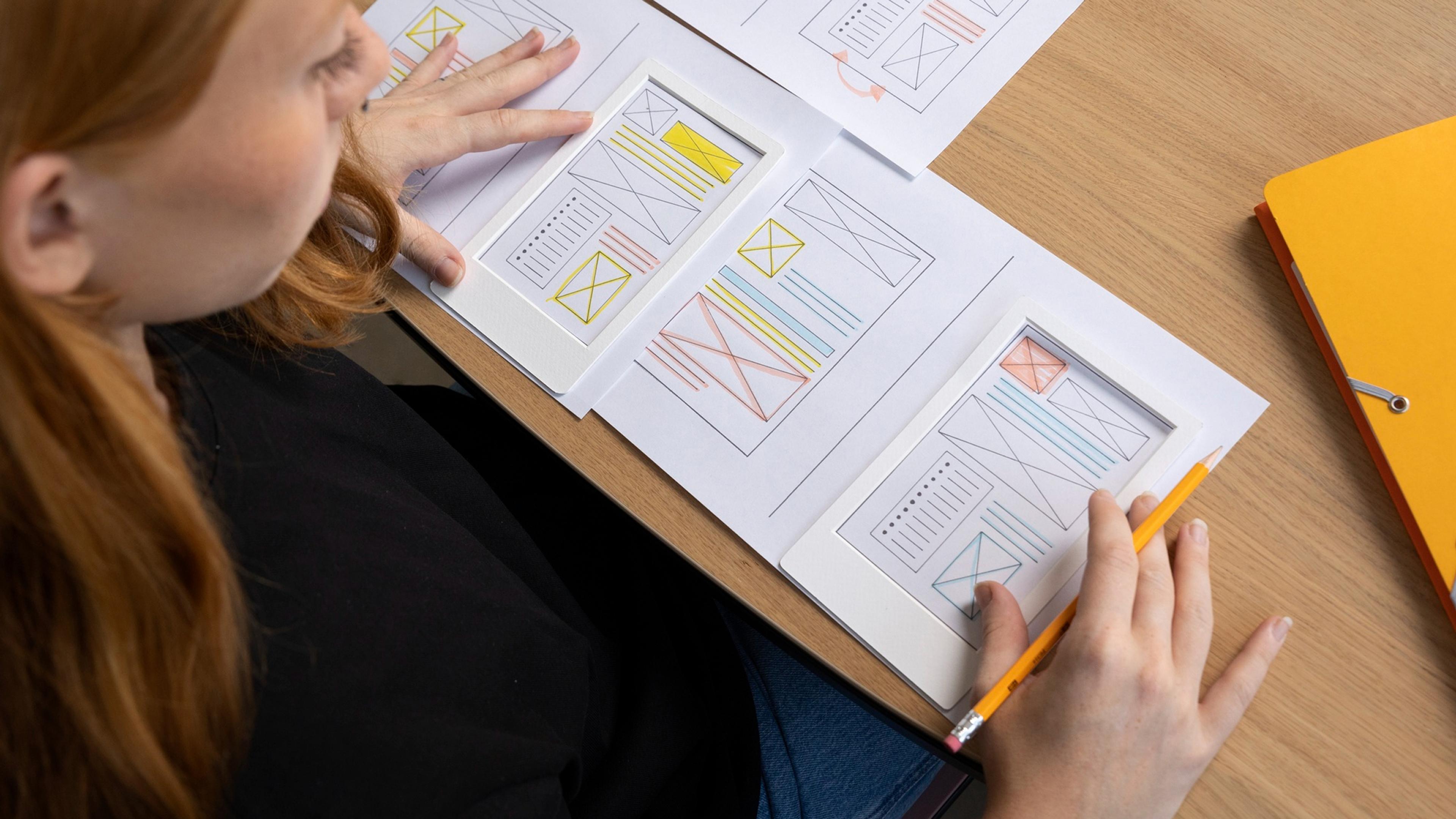A woman is looking at wireframe designs, with a yellow folder next to her on a wooden table
