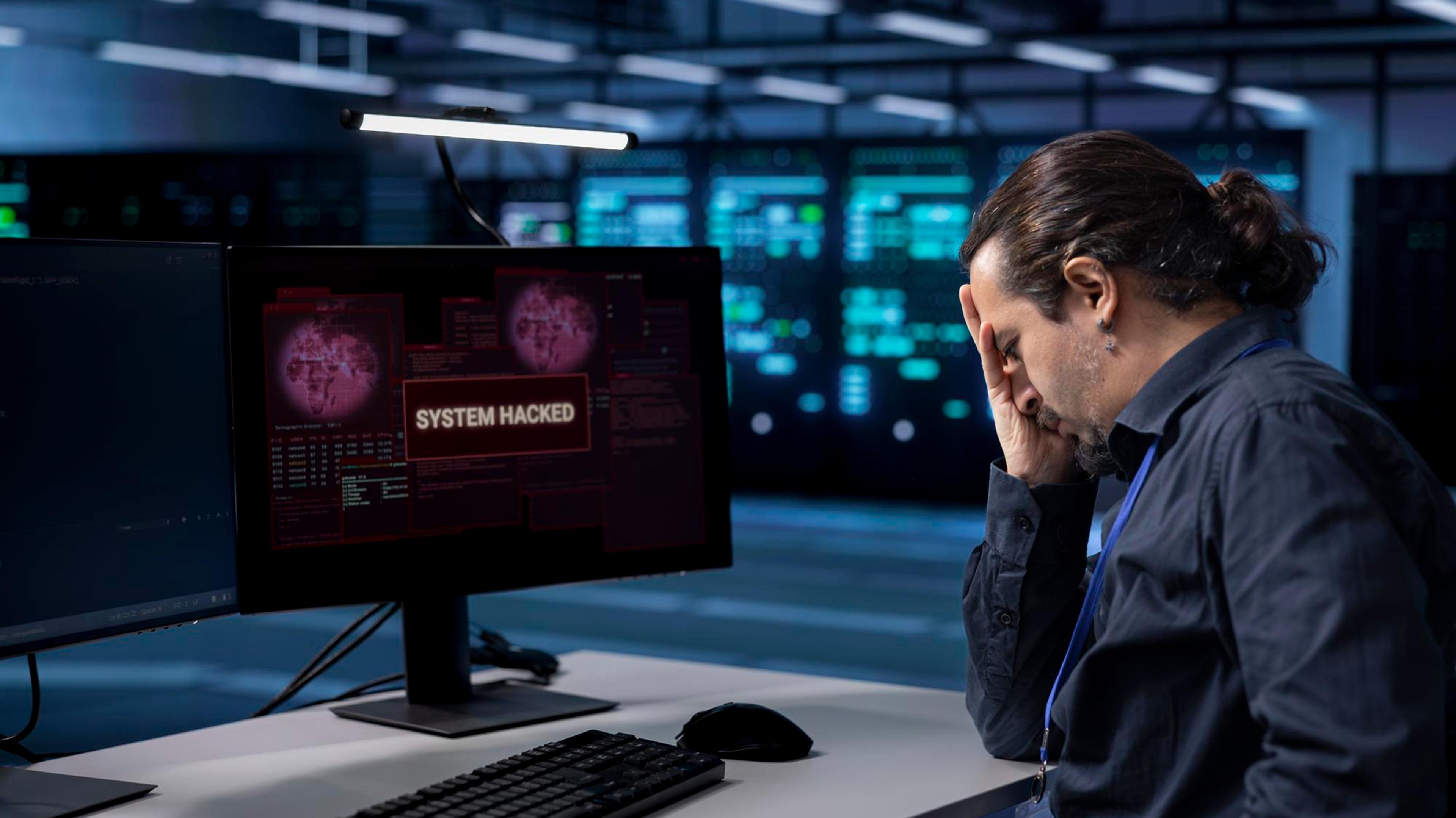 Обеспокоенный мужчина с рукой на лице перед экраном компьютера с надписью «Система взломана» в компьютерном центре.