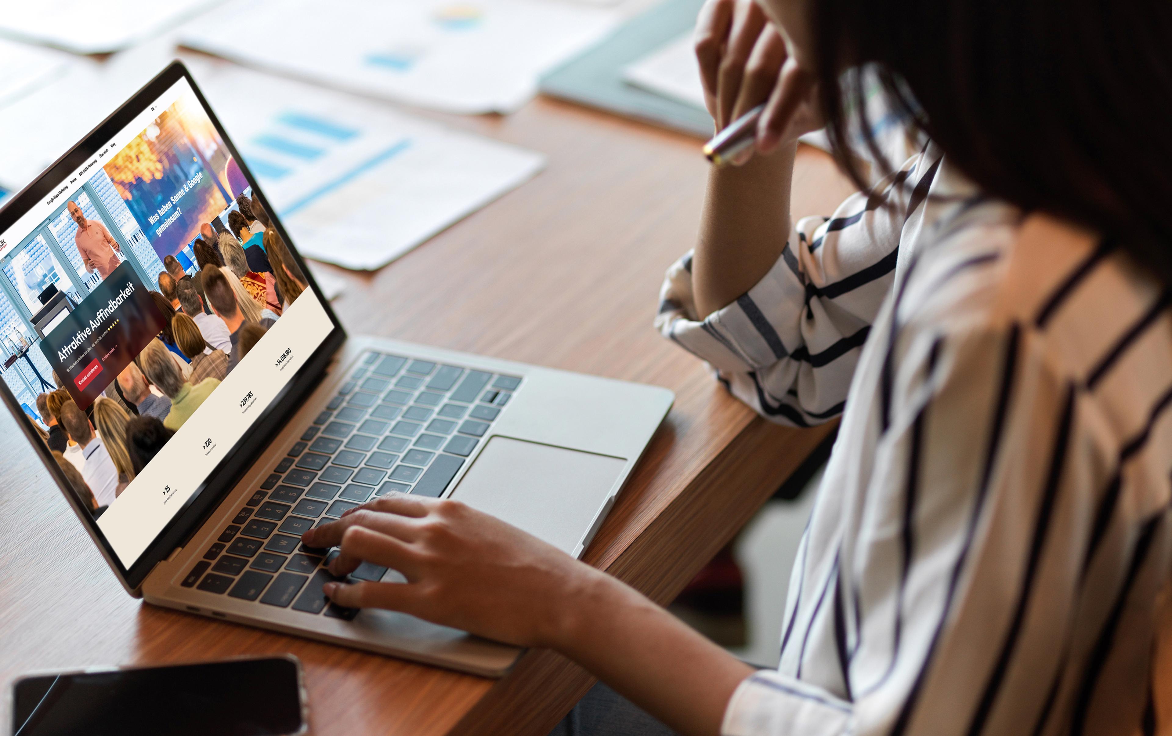 Eine Person in gestreiftem Hemd sitzt an einem Holzschreibtisch und betrachtet eine SEO-Seminar-Webseite auf ihrem Laptop