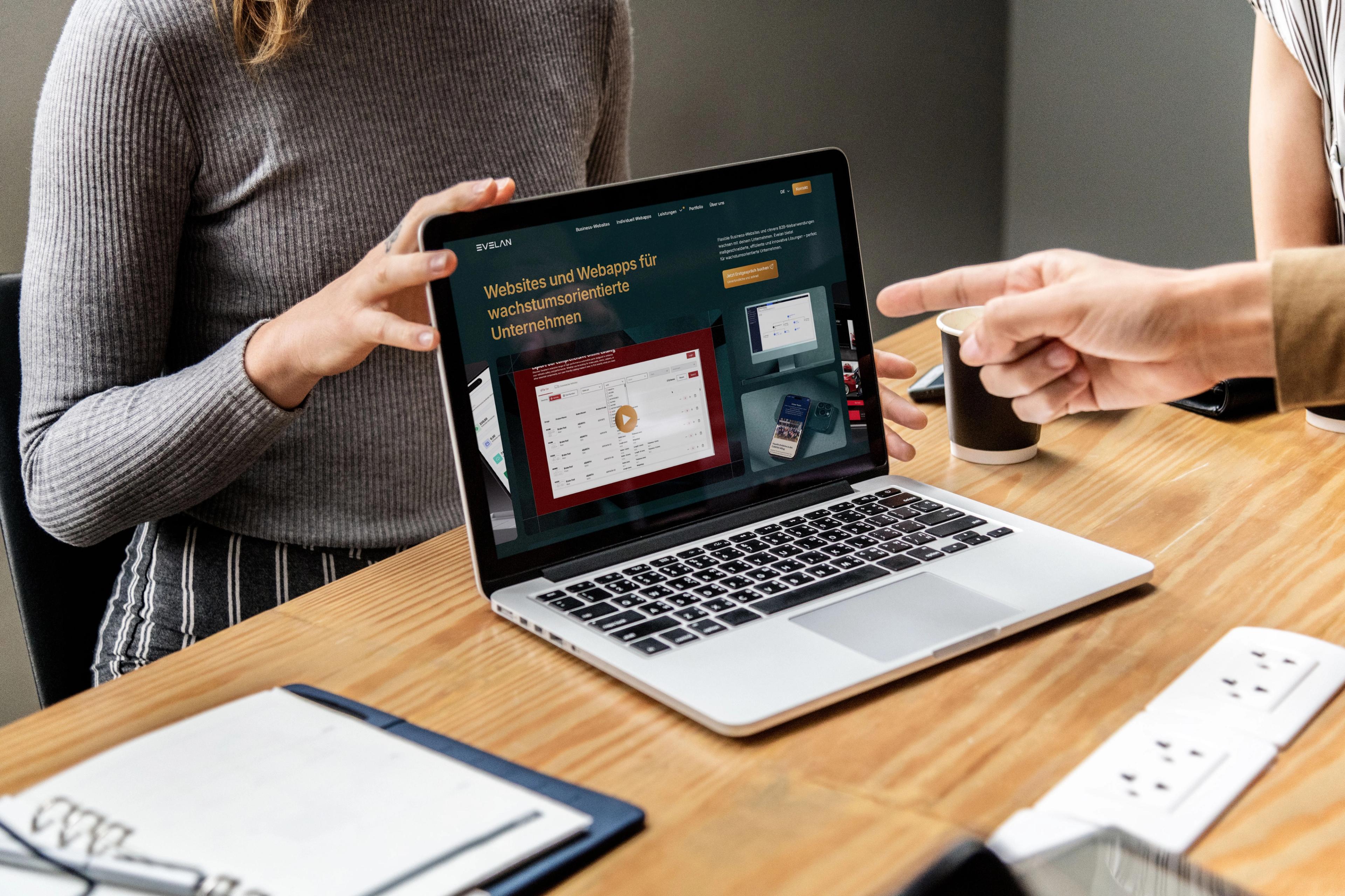 Eine Frau hält einen aufgeklappten Laptop mit der Webseite der Agentur Evelan, während ein Kollege mit dem Finger auf den Bildschirm zeigt
