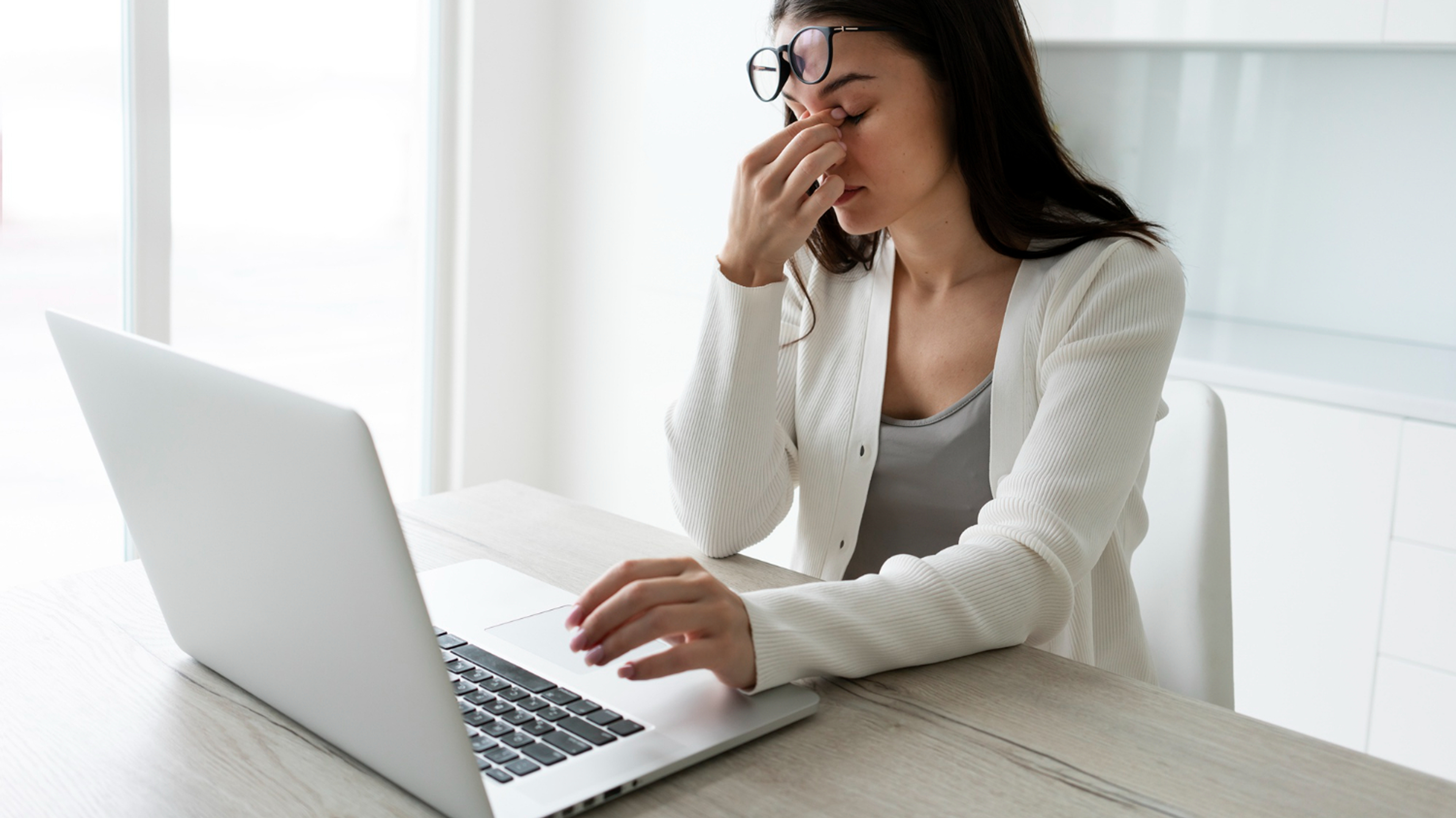 Eine junge Frau sitzt an einem Holztisch vor einem Laptop in einem hellen Raum und reibt sich erschöpft die Augen.