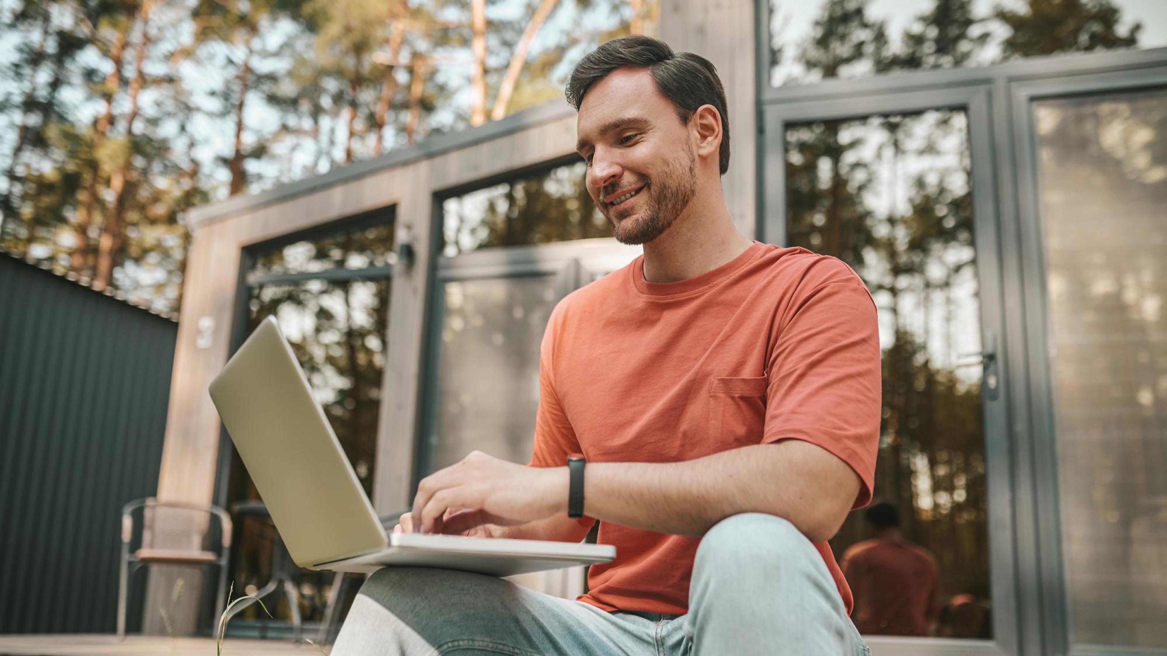 A man in a red T-shirt is working outdoors on a laptop. Behind him are glass windows and trees.