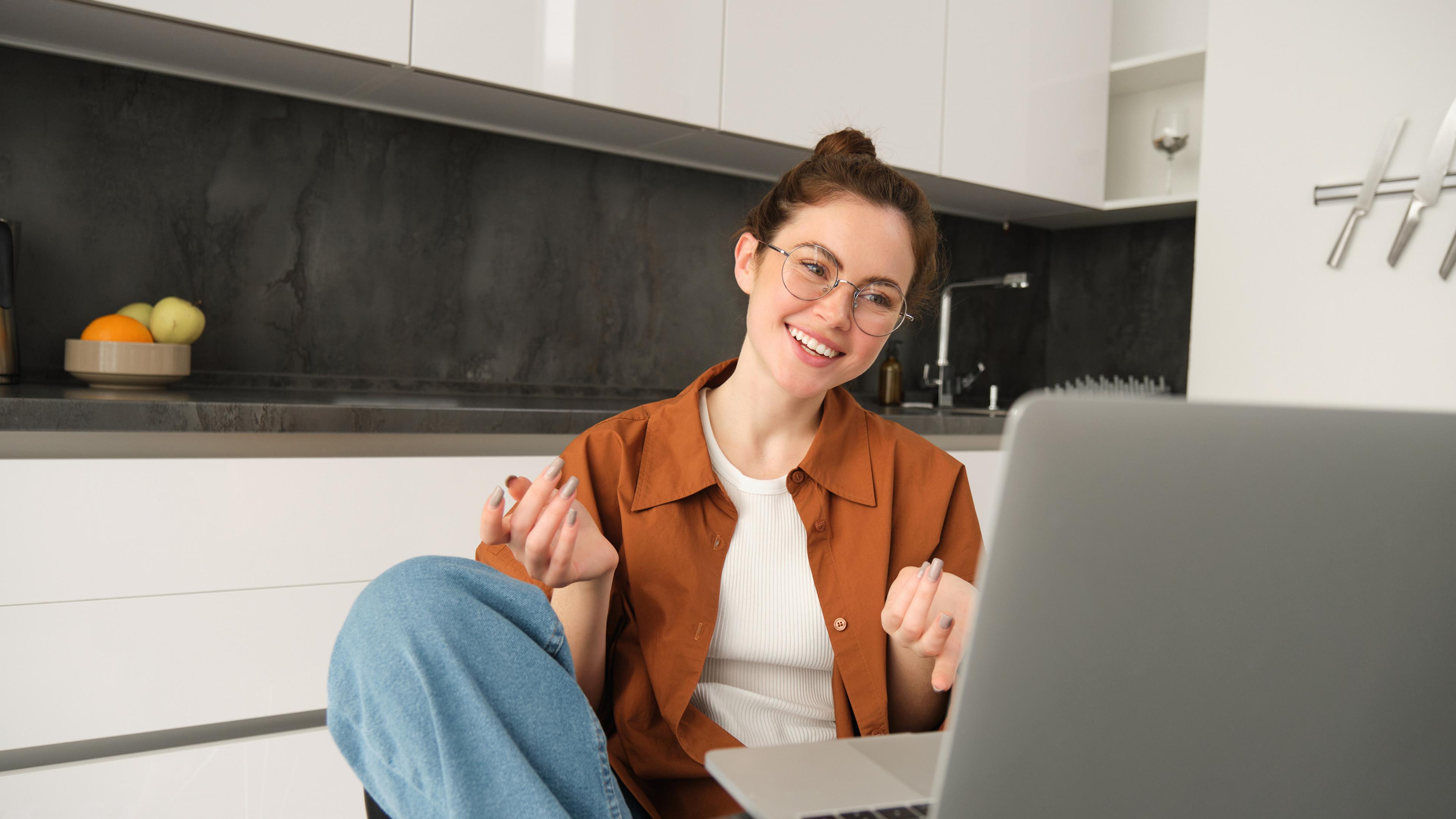 A woman wearing a brown blouse and glasses on a video call while working from home, with a laptop on the table in front of her.