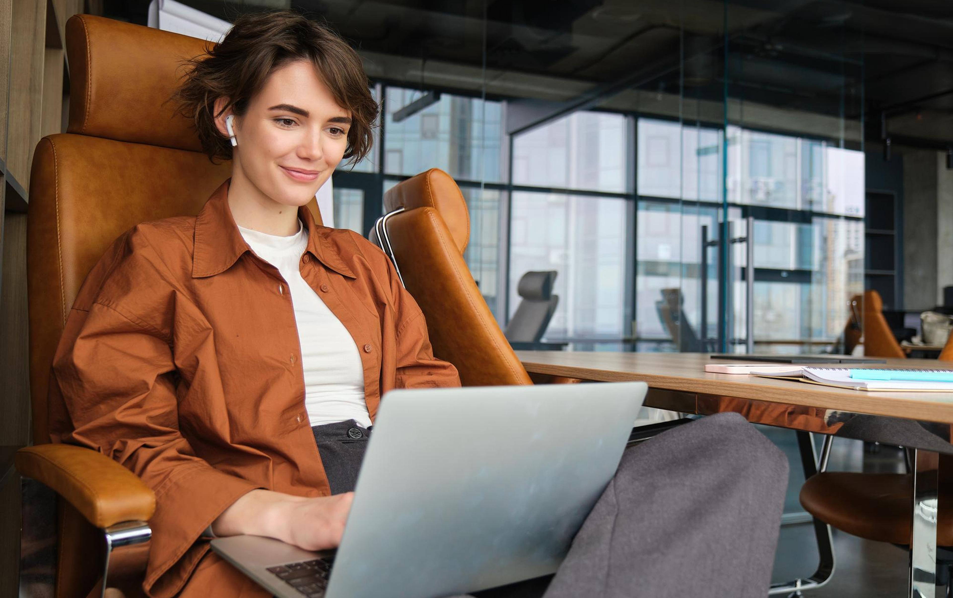 Женщина с короткими волосами в коричневой куртке сидит за ноутбуком в современном офисе.