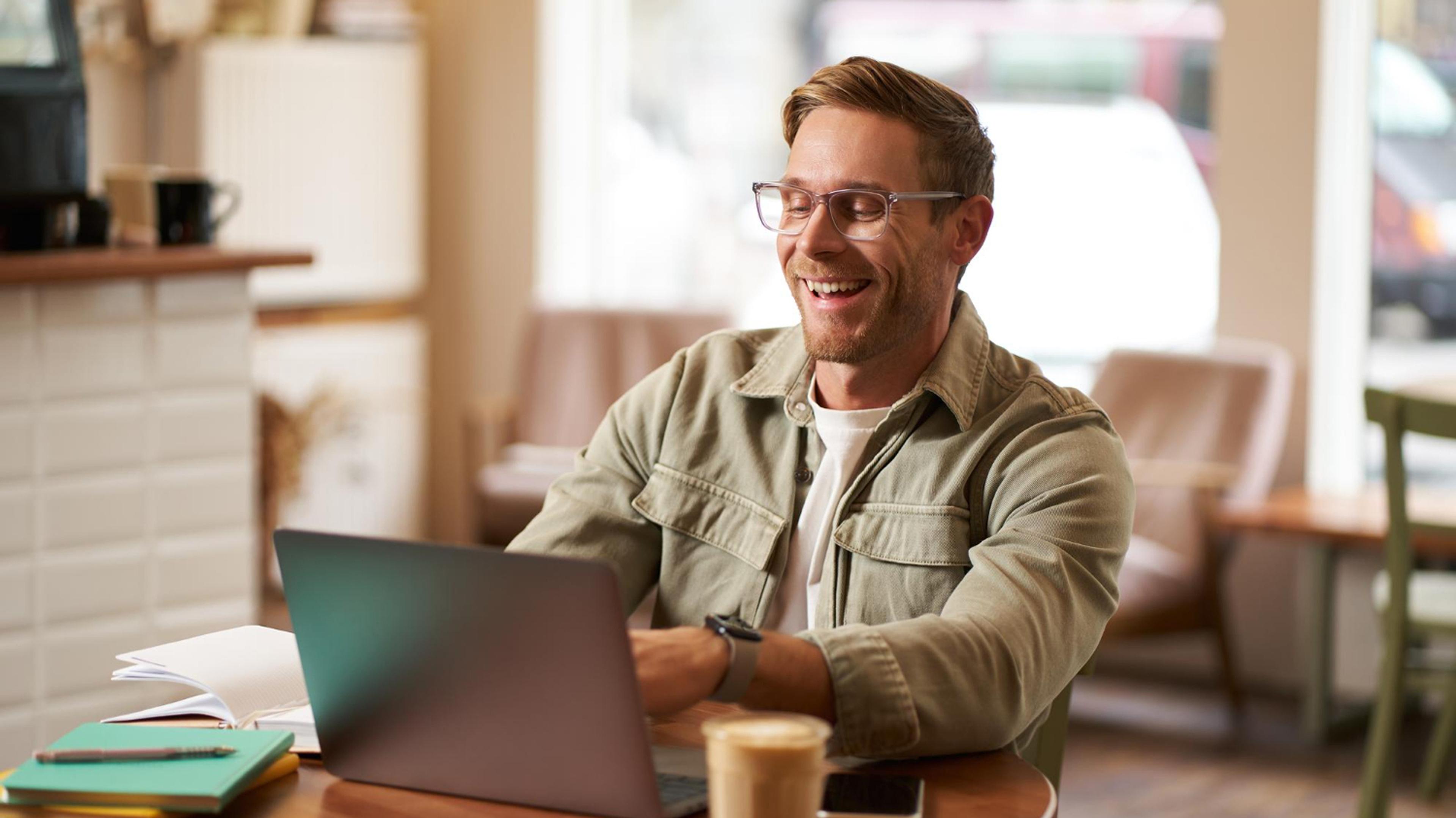 Mann mit Brille arbeitet lächelnd an einem Laptop im Café, Kaffee und Notizbuch auf dem Tisch