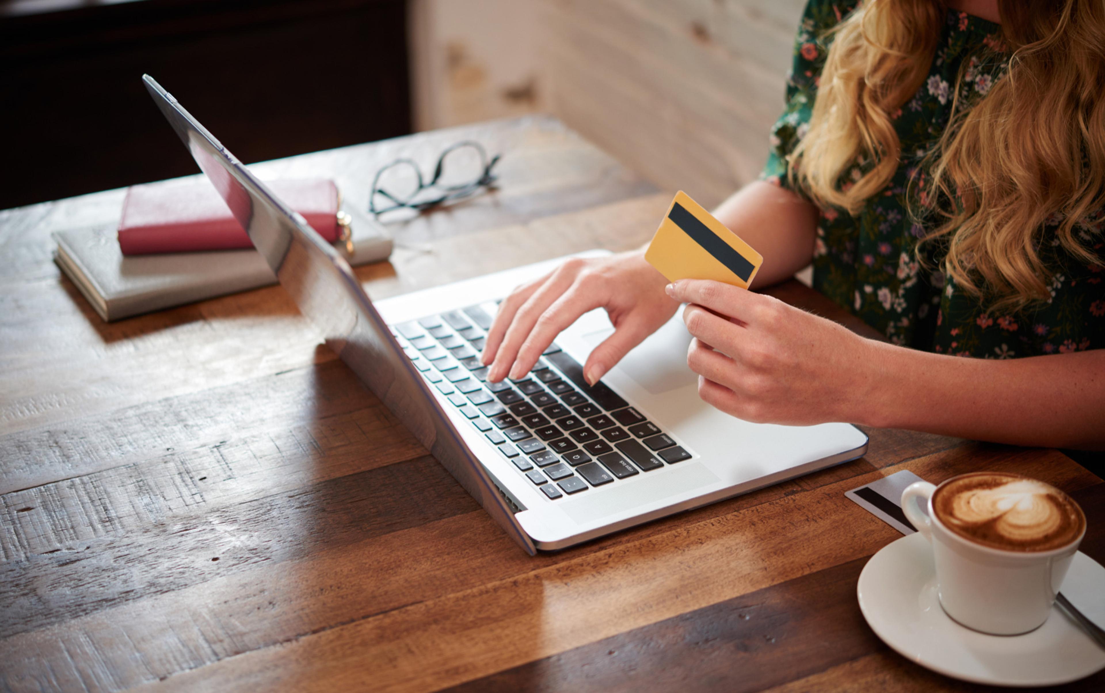Person tippt auf Laptop und hält Kreditkarte. Auf dem Tisch ist ein Kaffee.