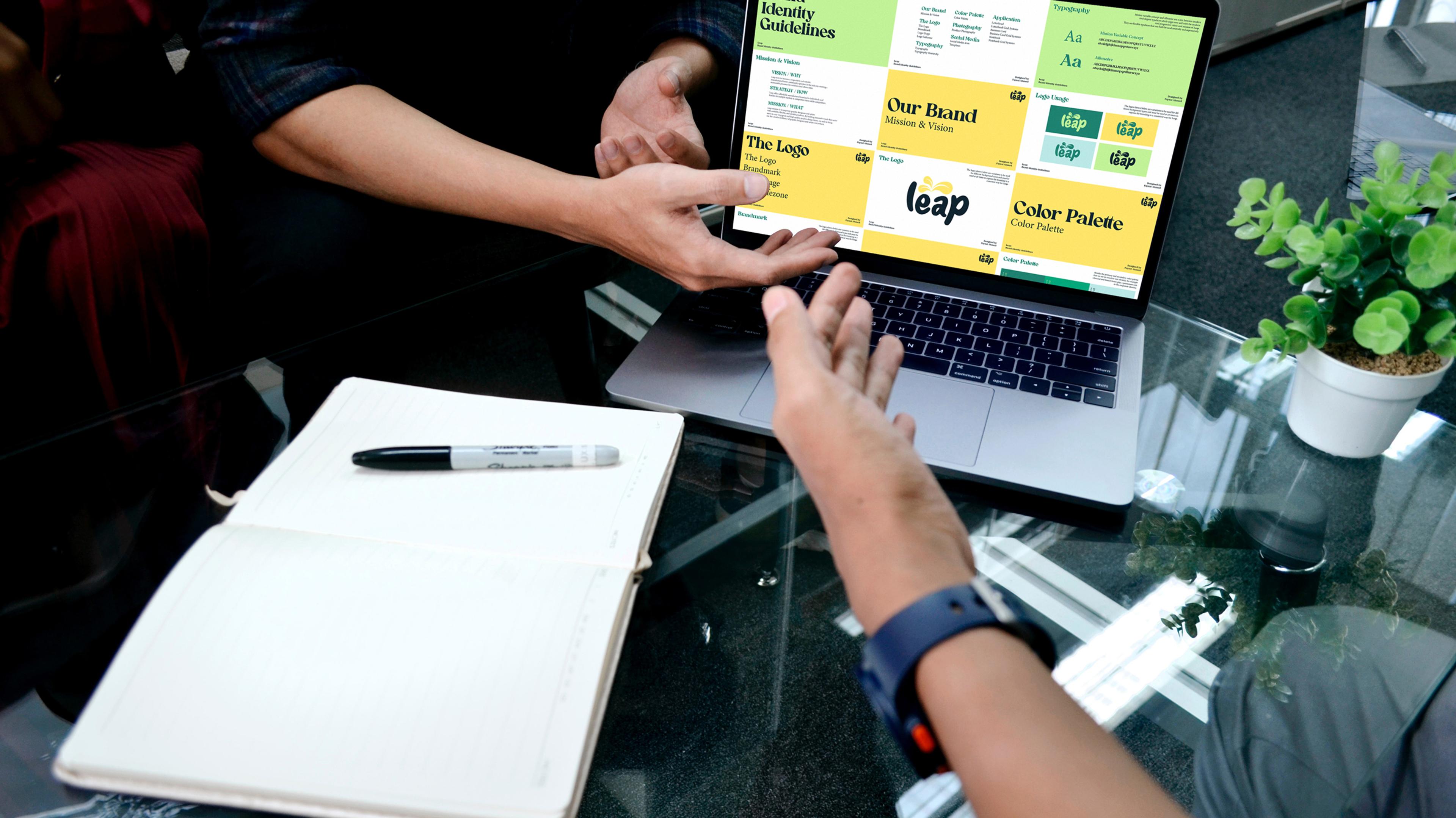 Hände gestikulieren vor einem Laptop mit Markenleitfaden. Am Tisch liegt ein Notizbuch.
