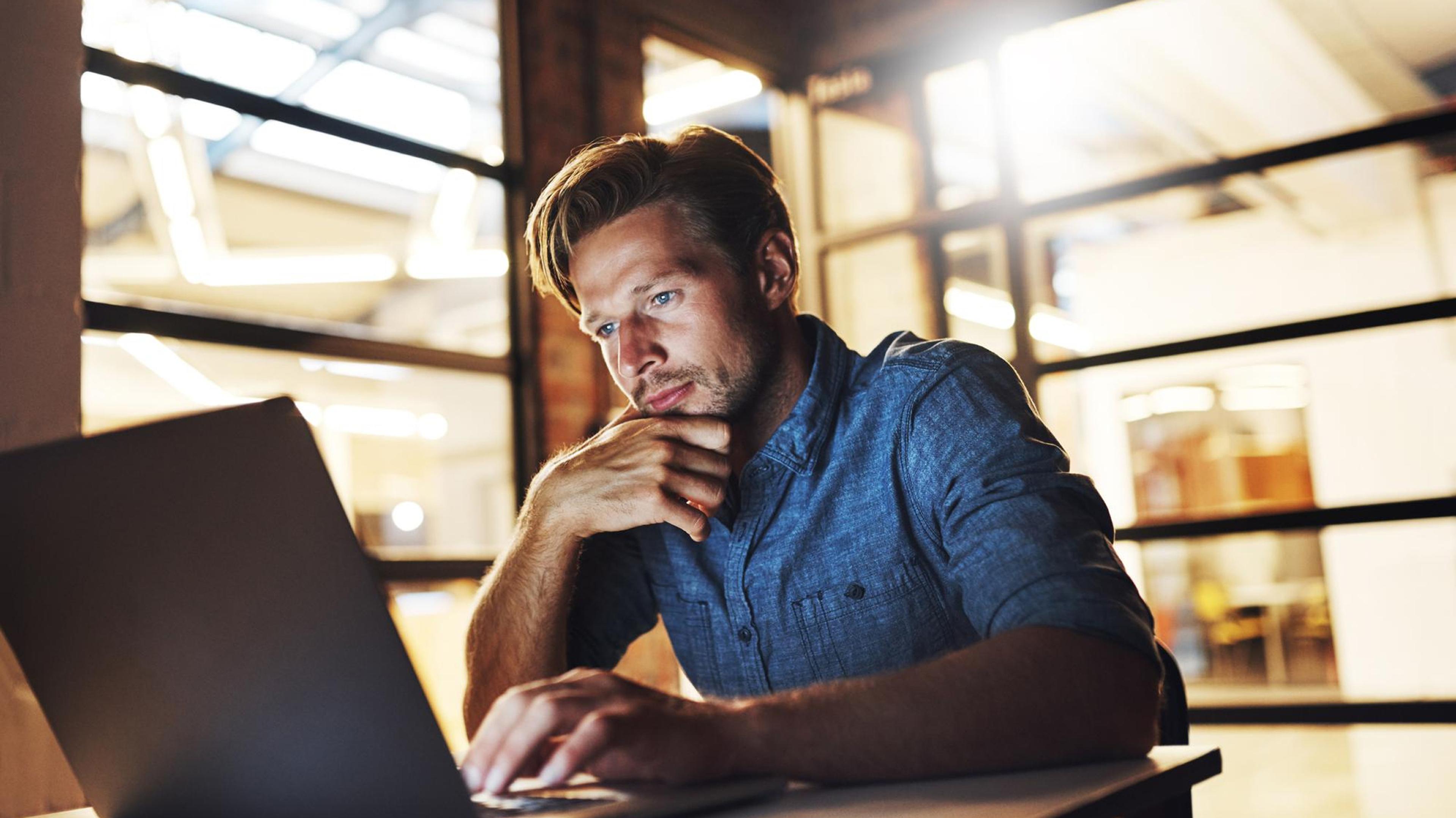 A man in a denim shirt works on his laptop in a modern office with glass walls