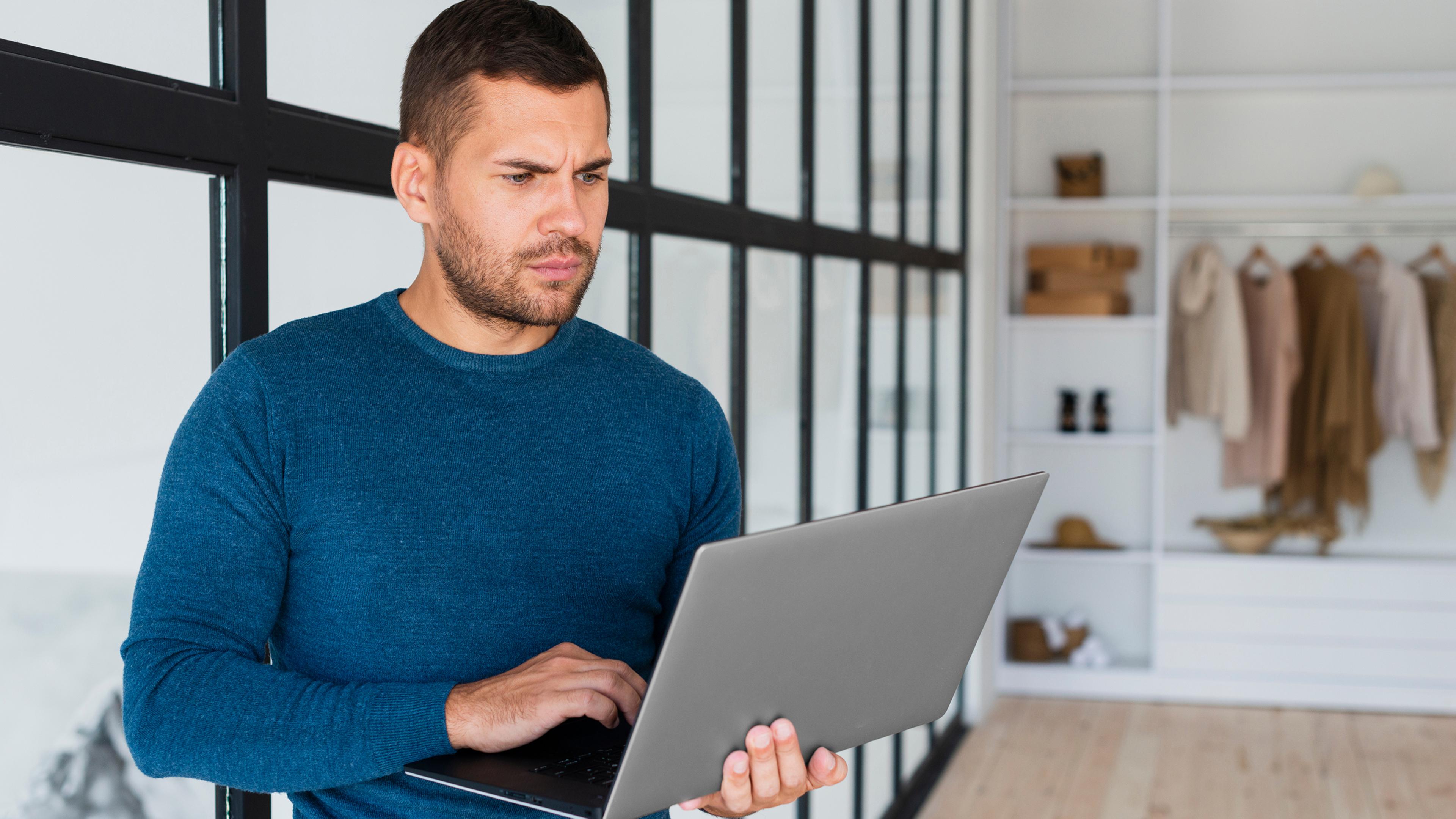 A man in a blue sweater holding a laptop in a modern office with clothes in the background