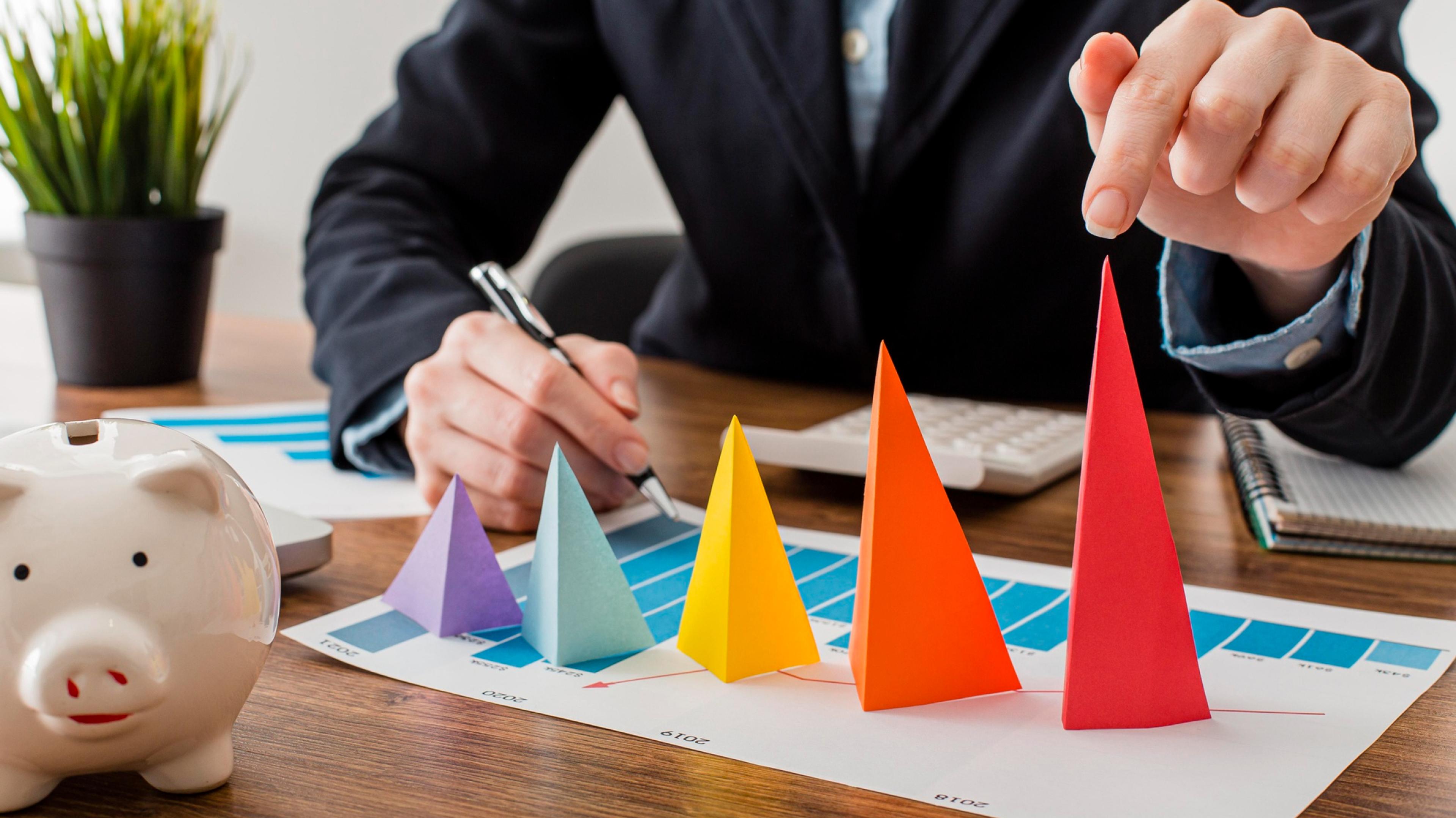 Eine Nahaufnahme von bunten Papierpyramiden auf einem gedruckten Diagramm, links ein weißes Sparschwein und rechts eine Hand in Business-Kleidung, die auf die höchste rote Pyramide zeigt.