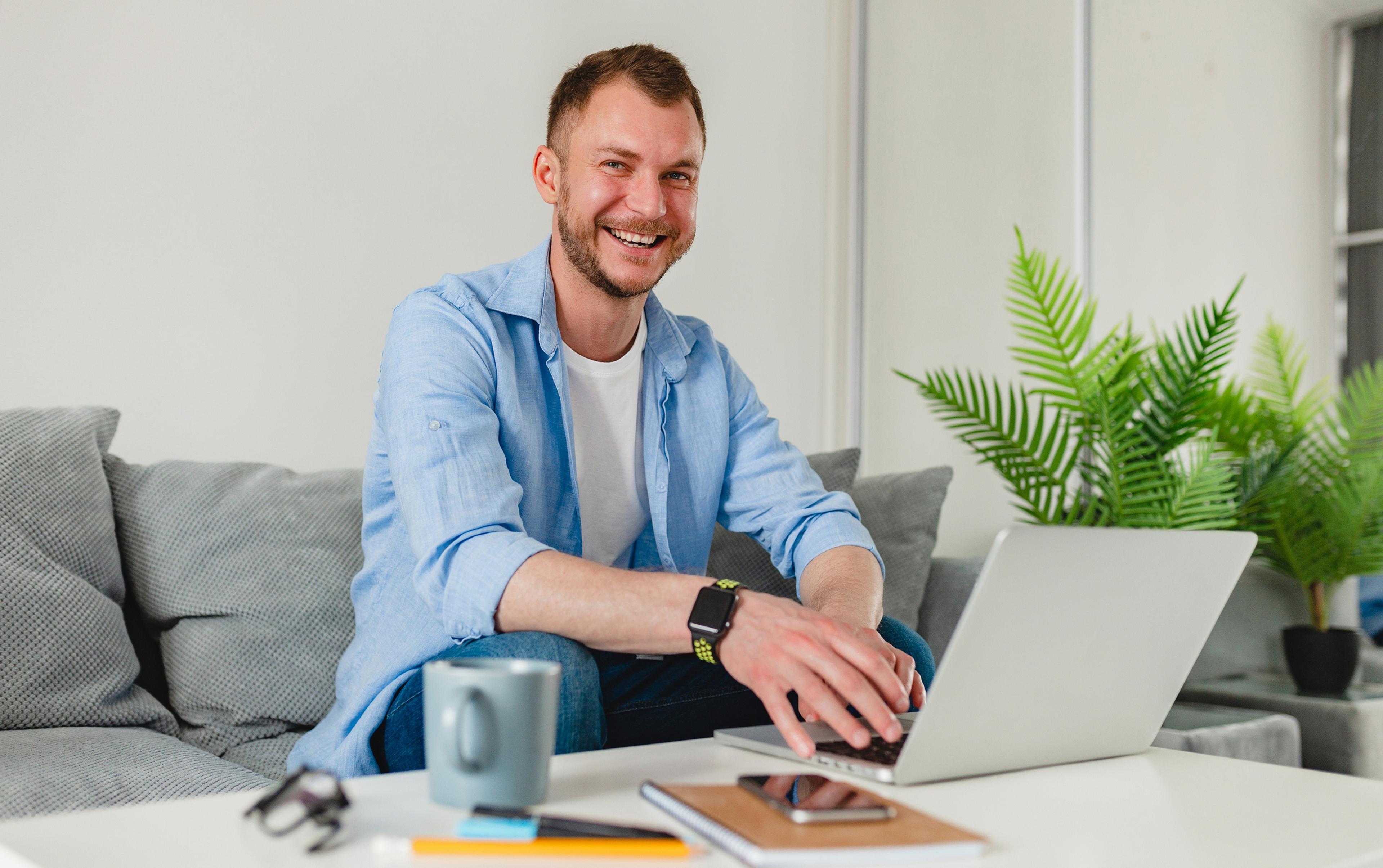 Lächelnder Mann mit hellem Hemd am Laptop auf Sofa, Pflanzen im Hintergrund