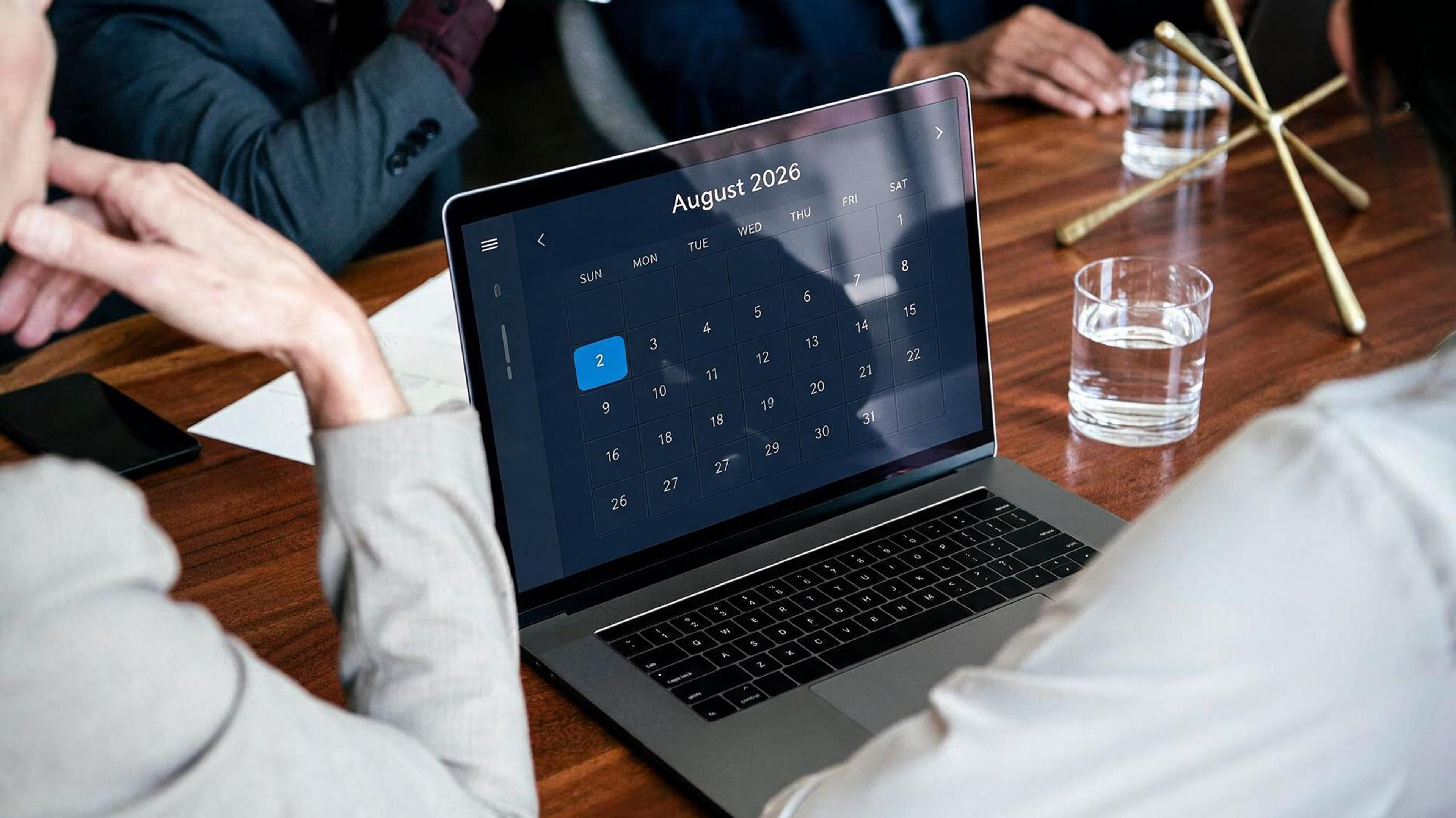 Bærbar datamaskin med kalender for august 2026 under et møte på et trebord