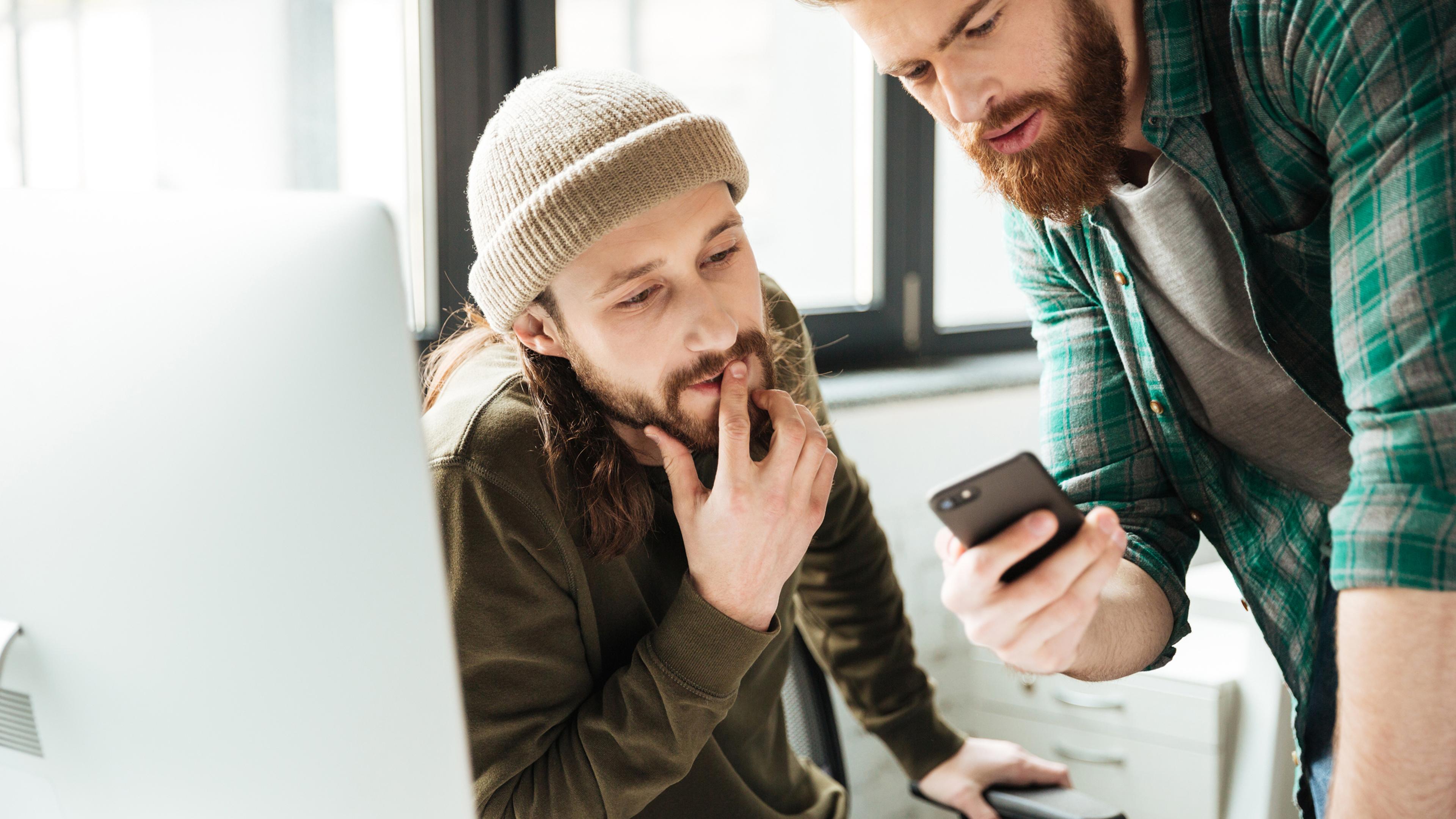 Zwei Männer im Büro, einer mit Mütze, betrachten ein Smartphone gemeinsam.
