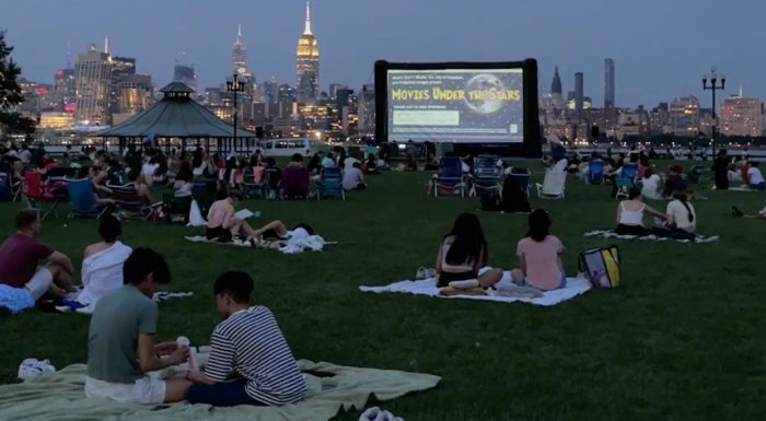 Movie screen on a lawn with people picnicing and Manhattan skyline in the background